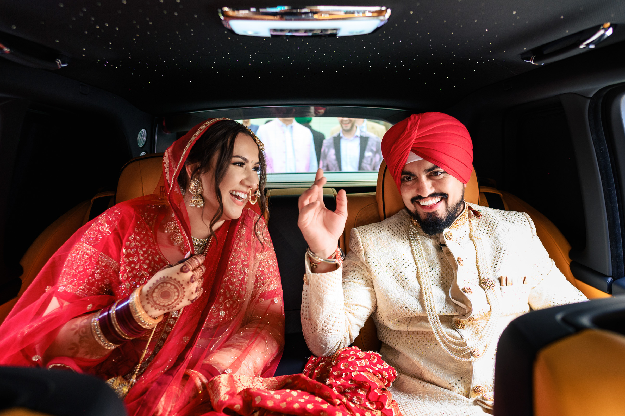 Bride and groom in traditional attire share a joyful moment inside a car.