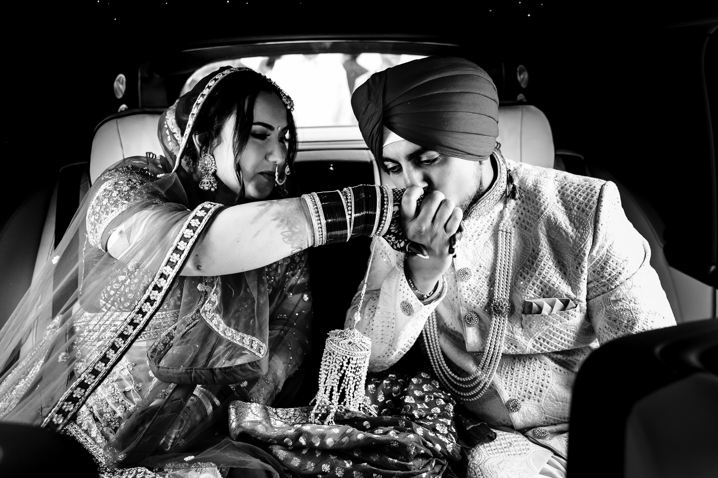 Bride and groom in traditional attire in a car; groom kisses bride's hand.