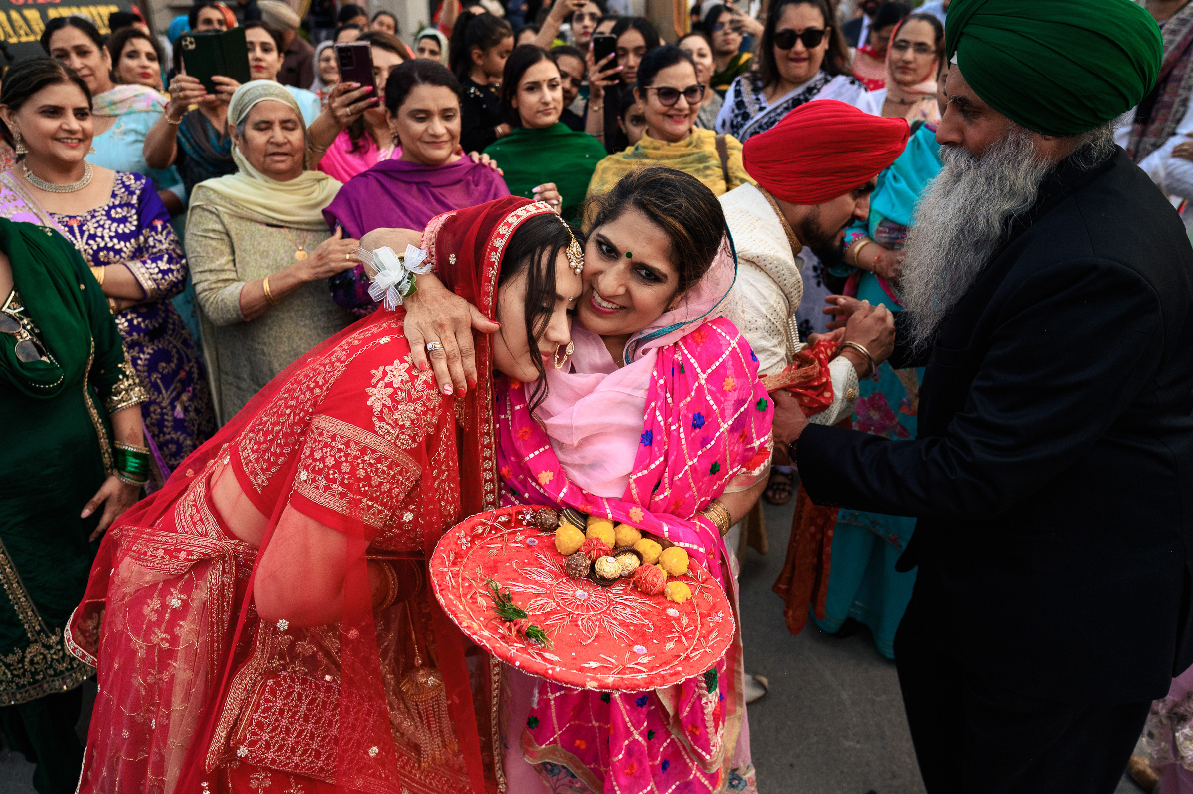 A bride in red is hugged by a woman holding a decorated plate, surrounded by a festive crowd.