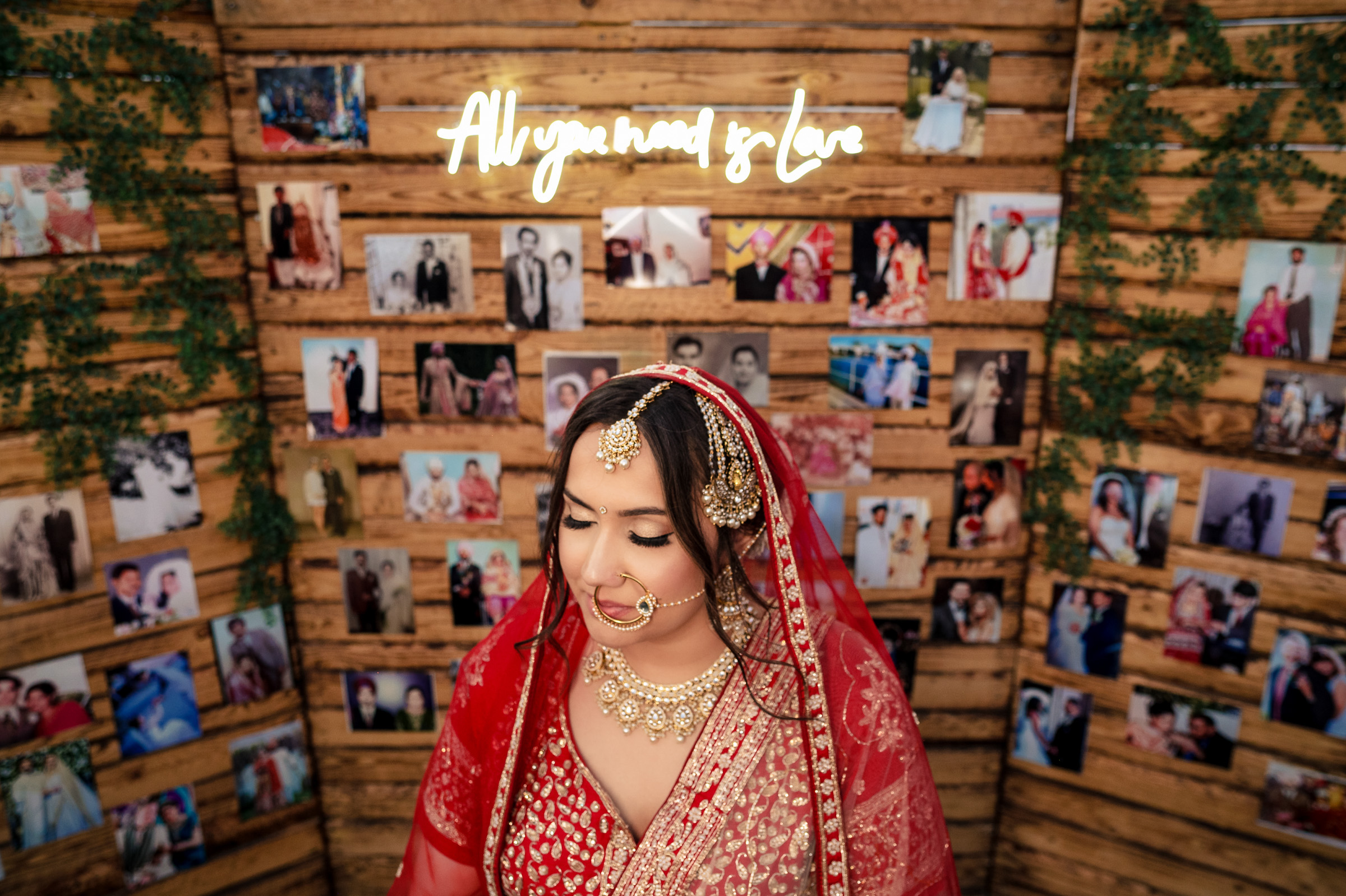 Bride in red attire stands before a wall with photos and a neon sign reading "All you need is love".