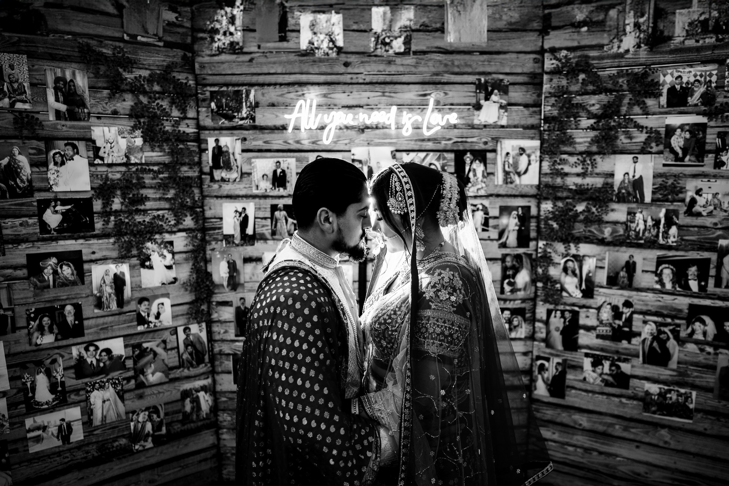 Couple in traditional attire touching foreheads in front of photo-covered wooden wall.