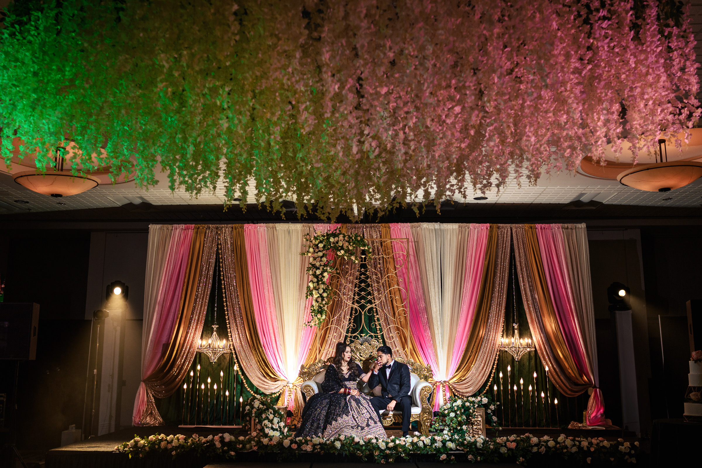 Elegant wedding stage with floral canopy, draped curtains, and a couple sitting together.