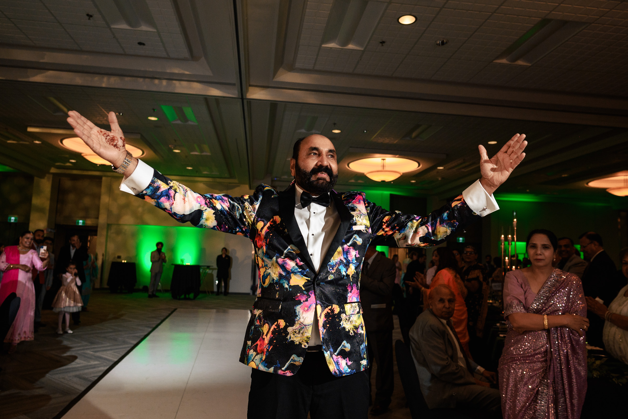 Person in a vibrant jacket gestures with open arms at an indoor event.