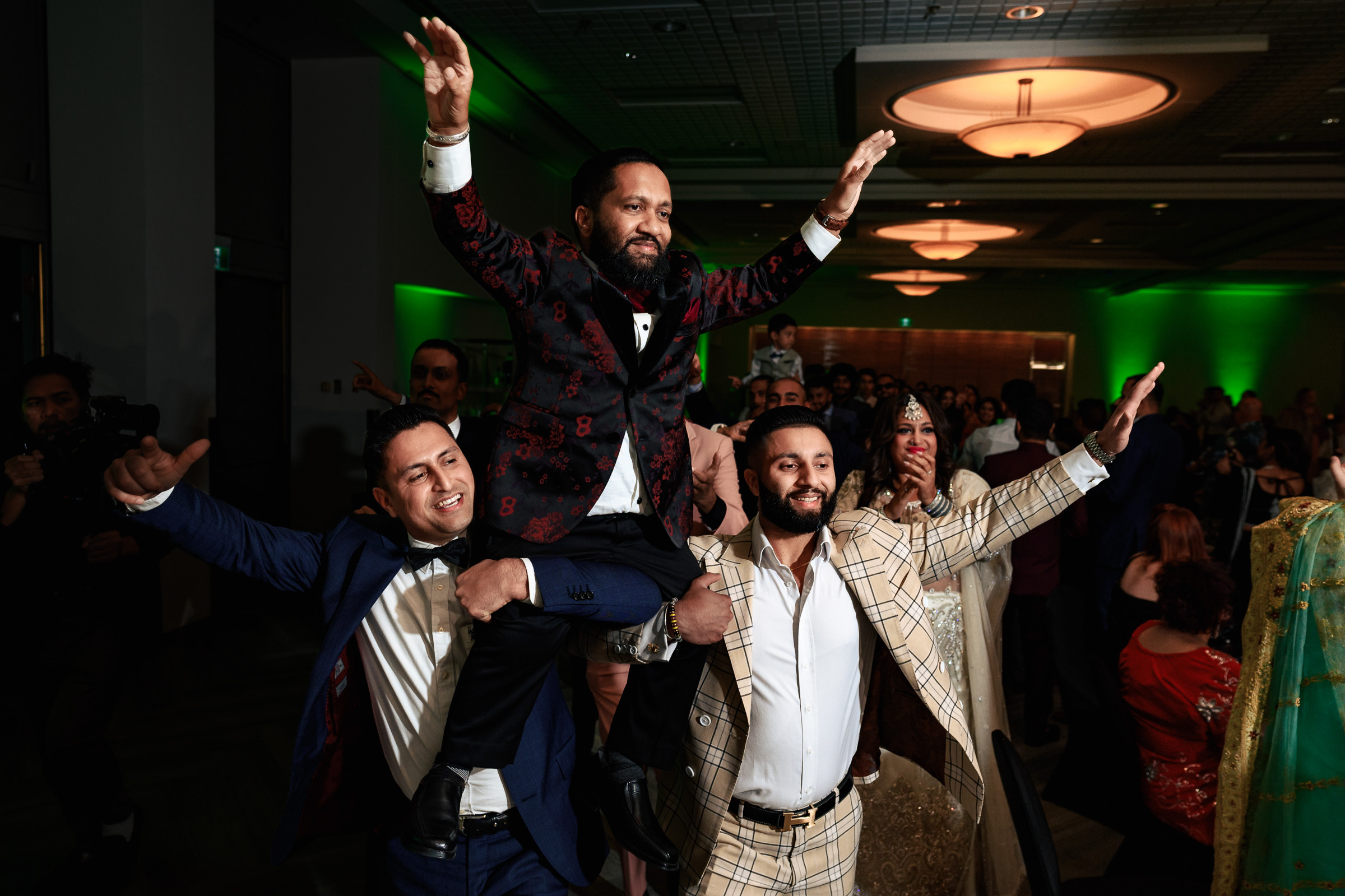 Three men celebrate at a party, carrying a man on their shoulders with arms raised.