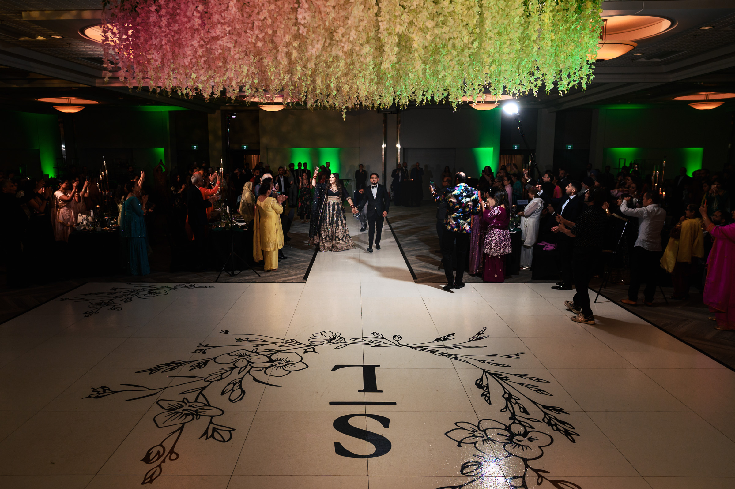 A couple walks onto the dance floor under hanging flowers at a formal event.
