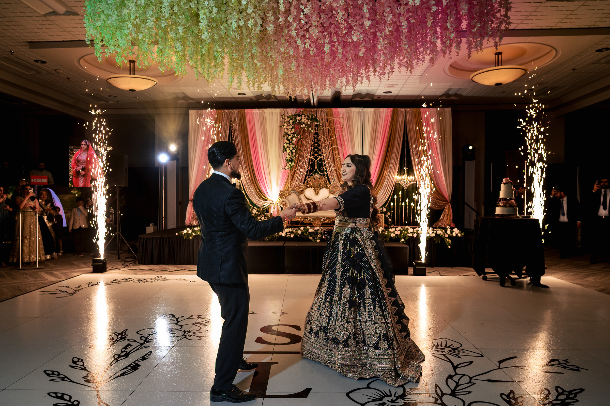 Couple dancing at a decorated ballroom event with floral ceiling and sparkler effects.