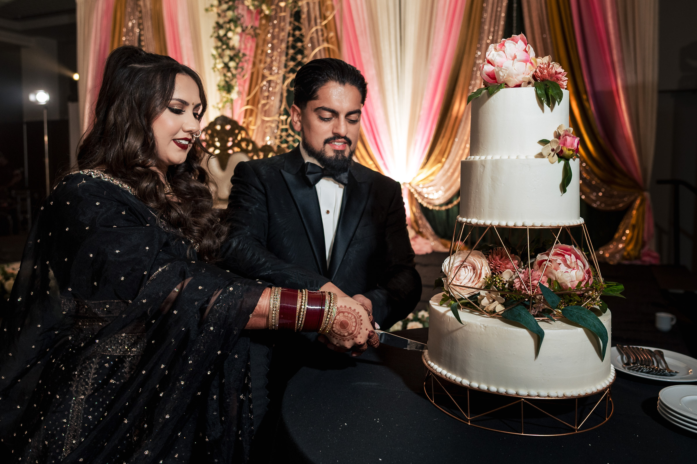 A couple cuts a tiered wedding cake adorned with pink flowers, set against a draped backdrop.