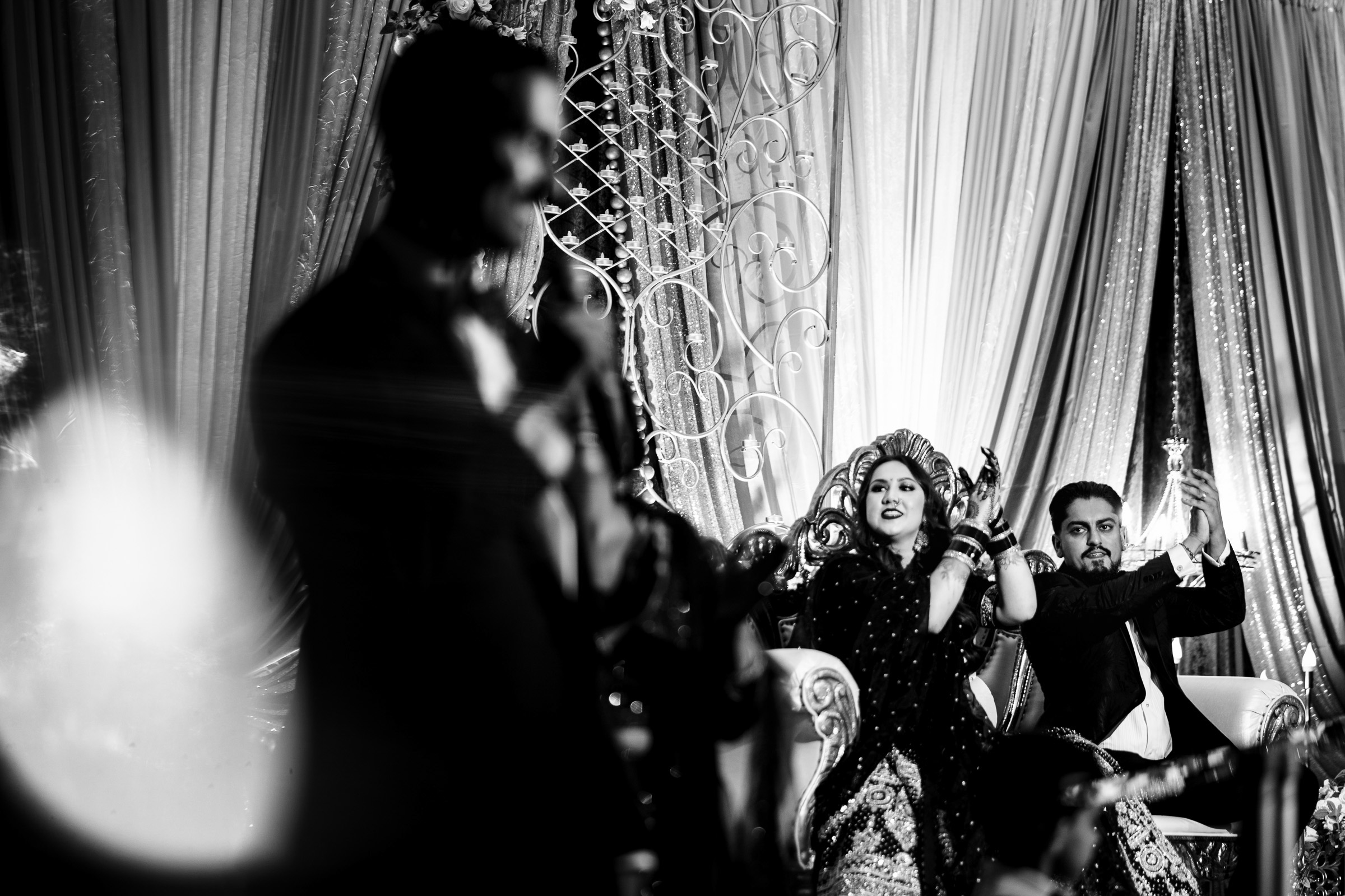 A couple sits on a decorated stage while a person stands in the foreground, out of focus.