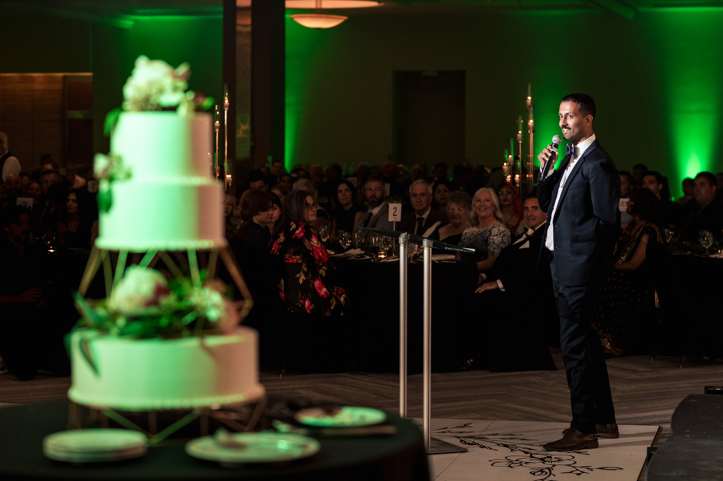 A man in a suit speaks at a formal event with a large tiered cake in the foreground.