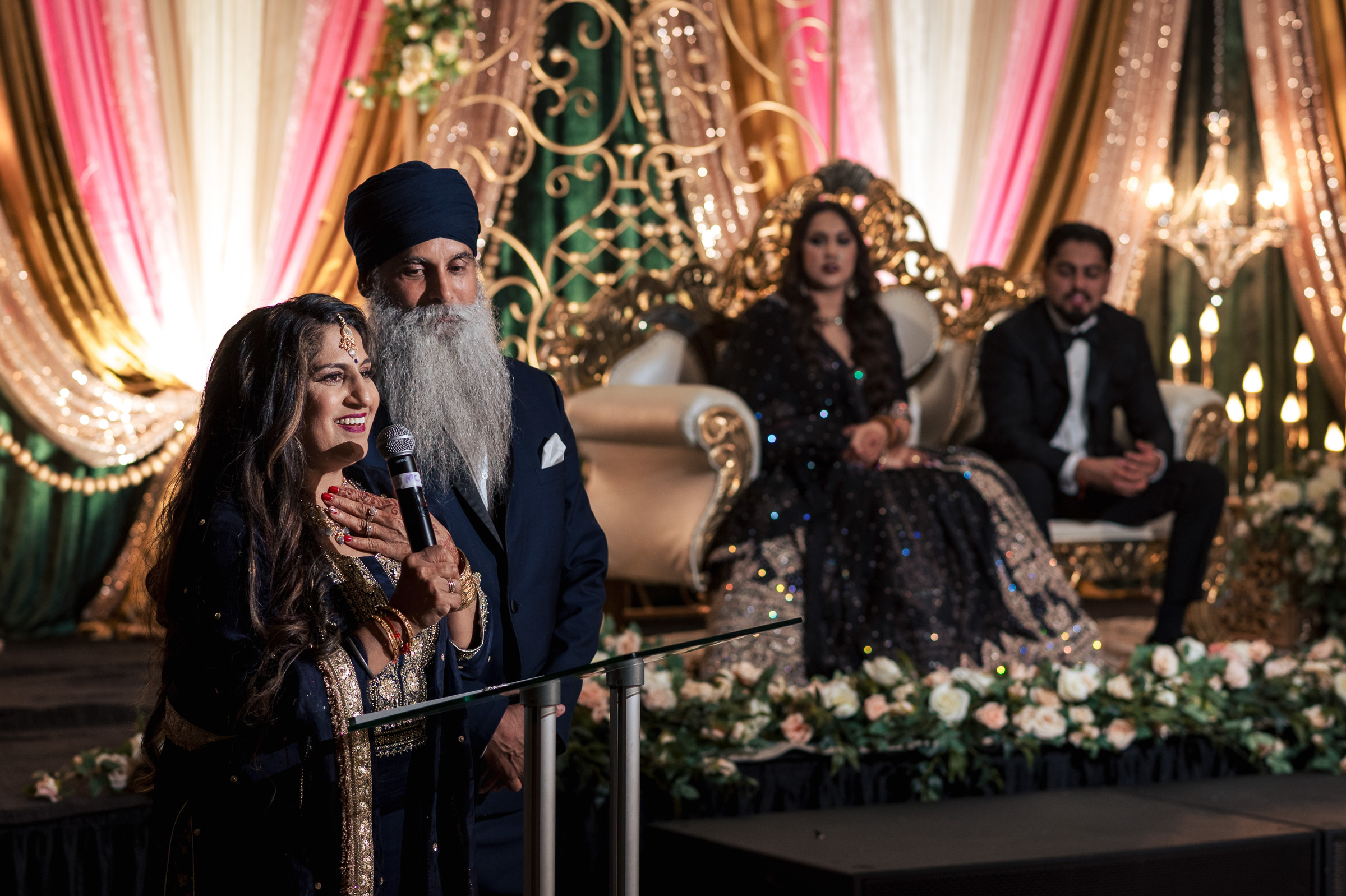 A woman speaks at a podium, two people sit on a decorated stage behind her.