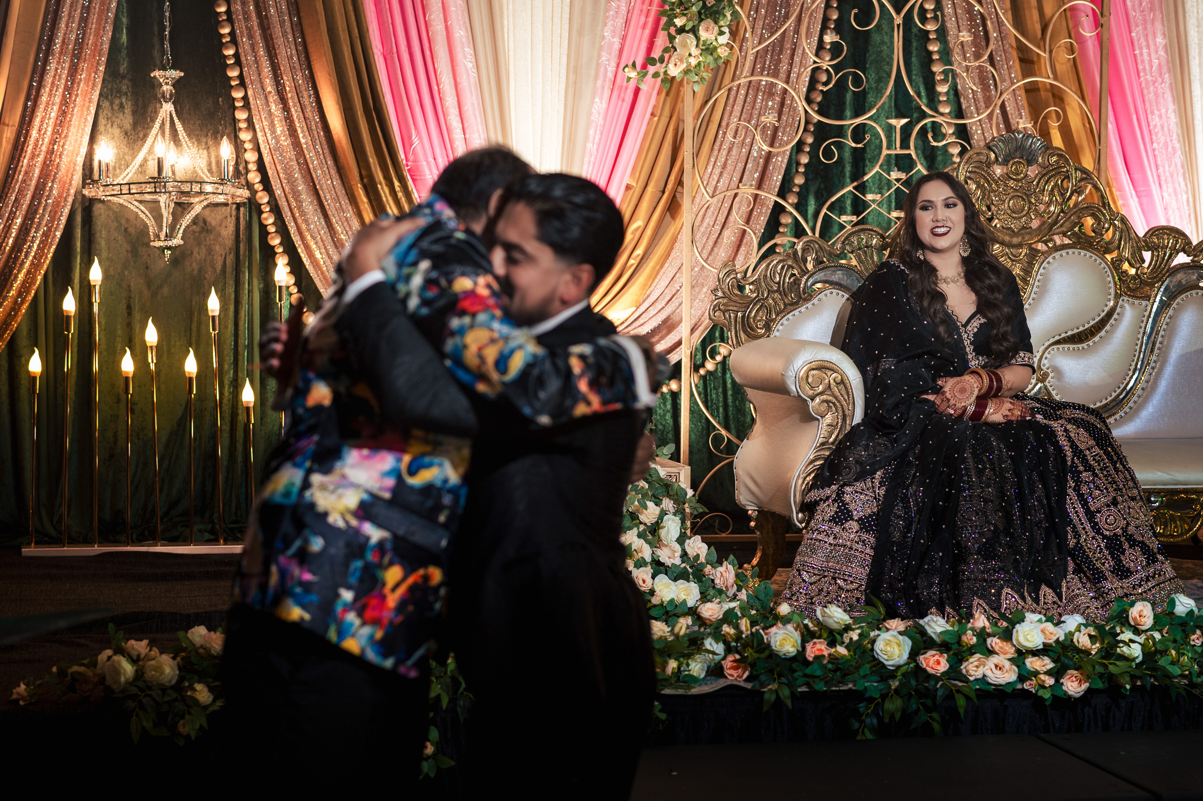 Two people hugging, woman in gown seated on ornate sofa, floral and draped backdrop.