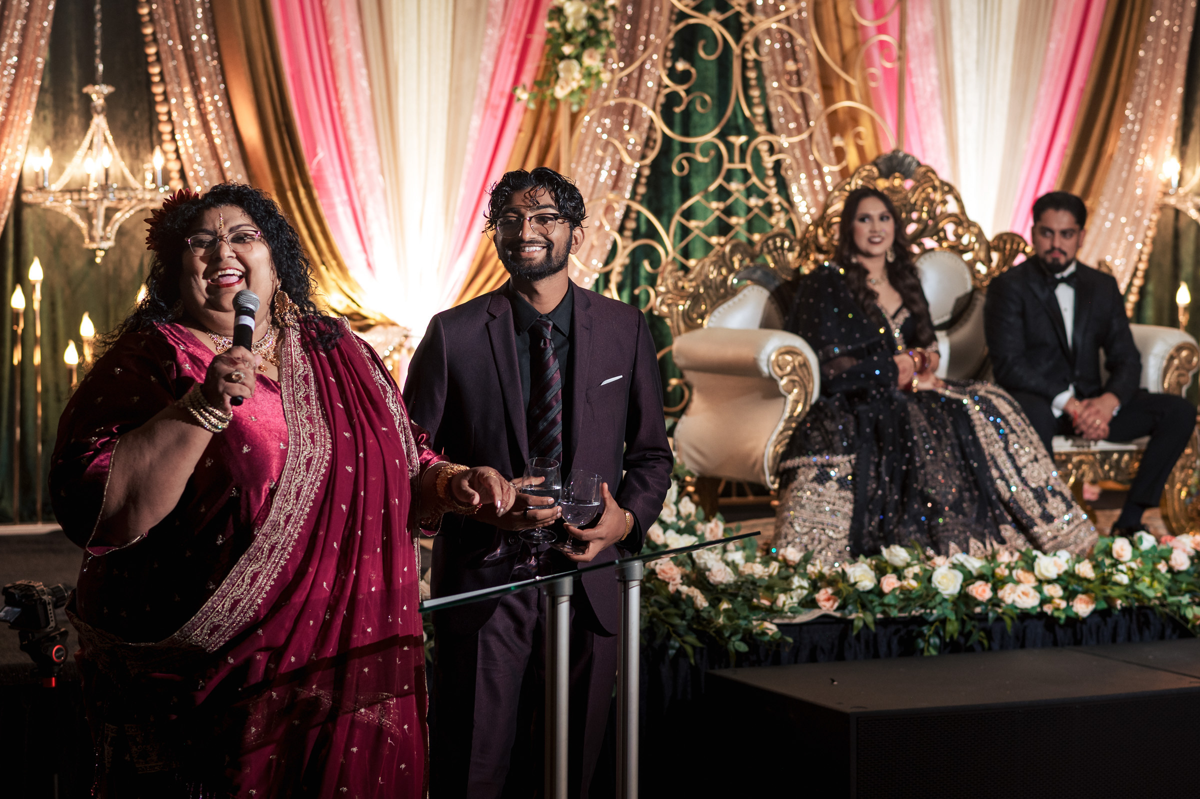 People giving a speech at a celebration with seated couple on a decorated stage.