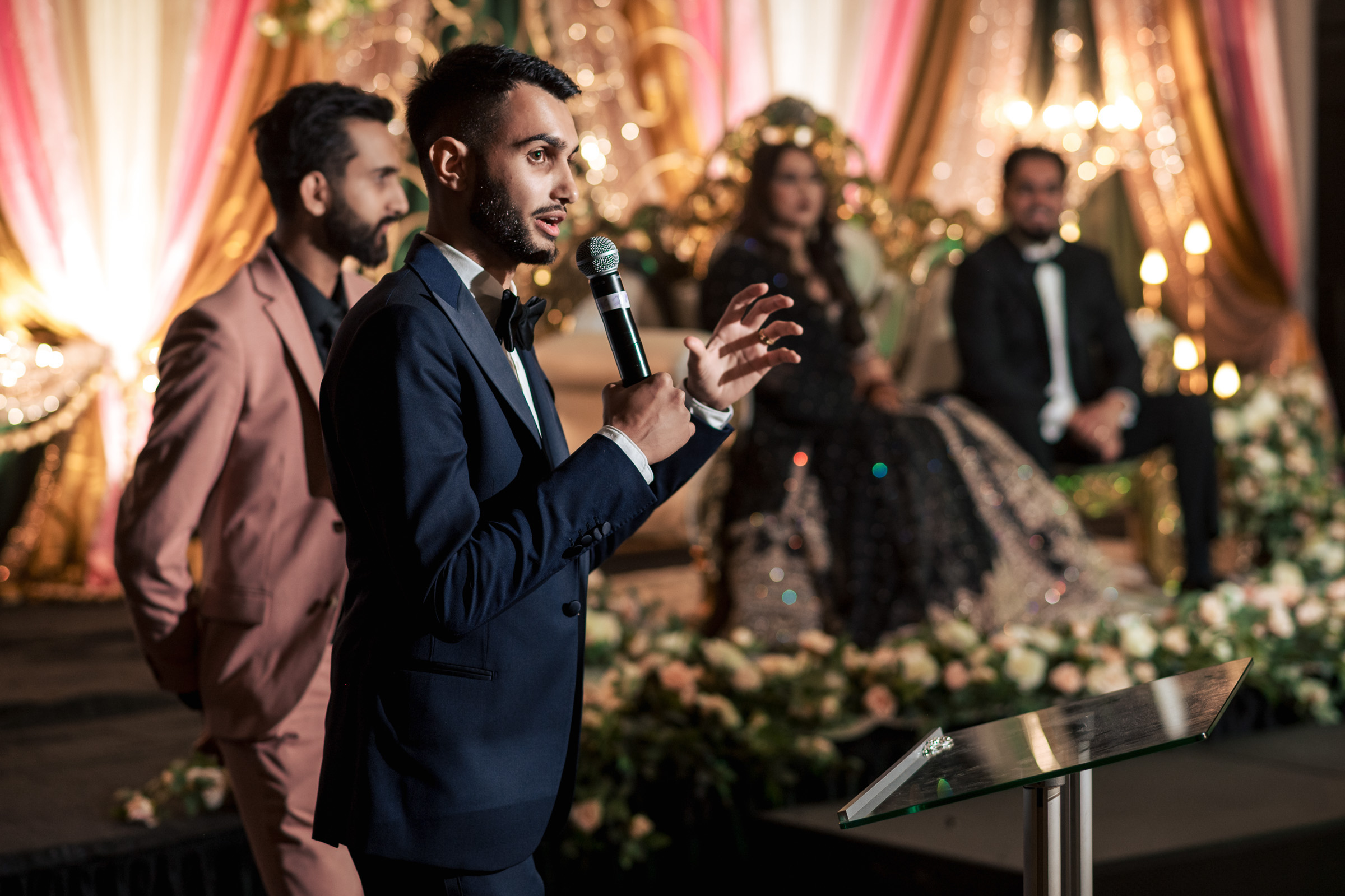 Man in suit speaking at an event, with seated guests in the background.
