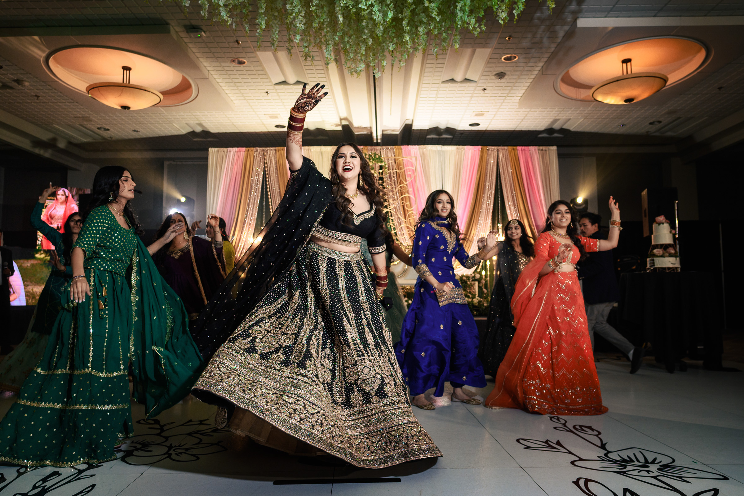 Women in colorful traditional dresses dance joyfully at an indoor celebration.