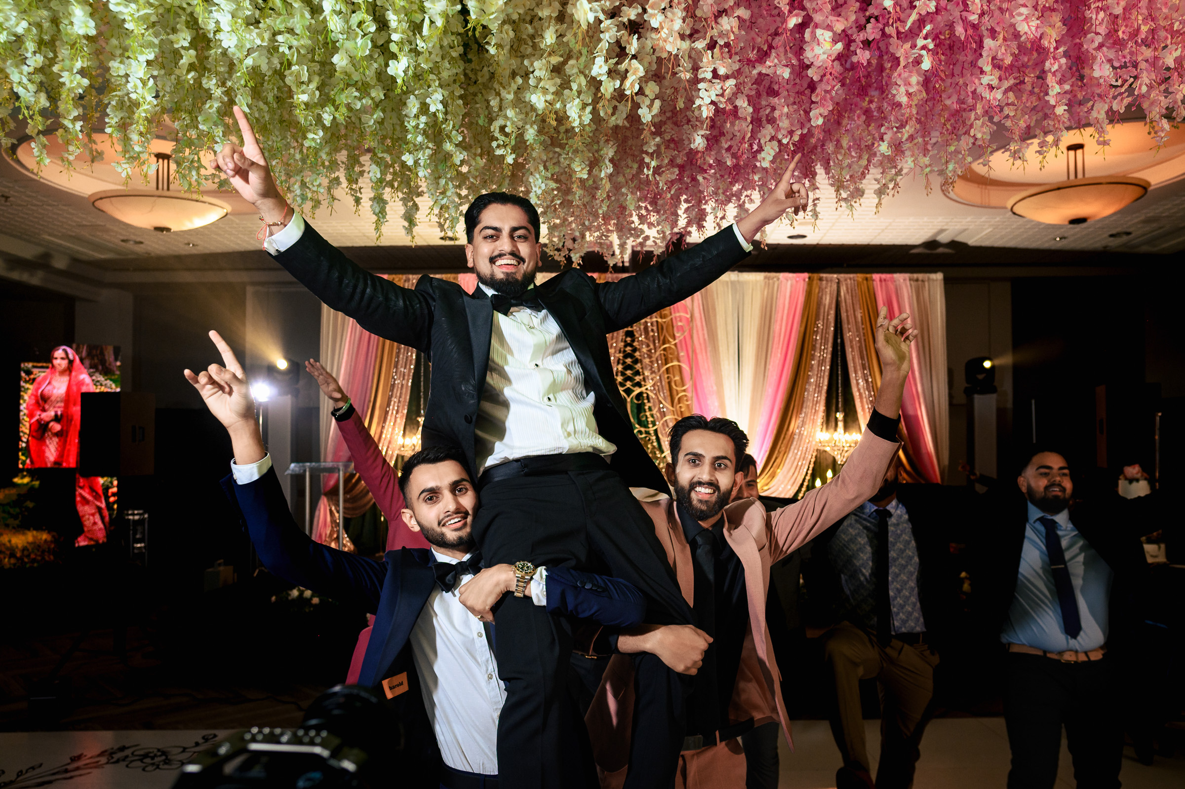 A group of men celebrating under colorful floral decorations at an indoor event.