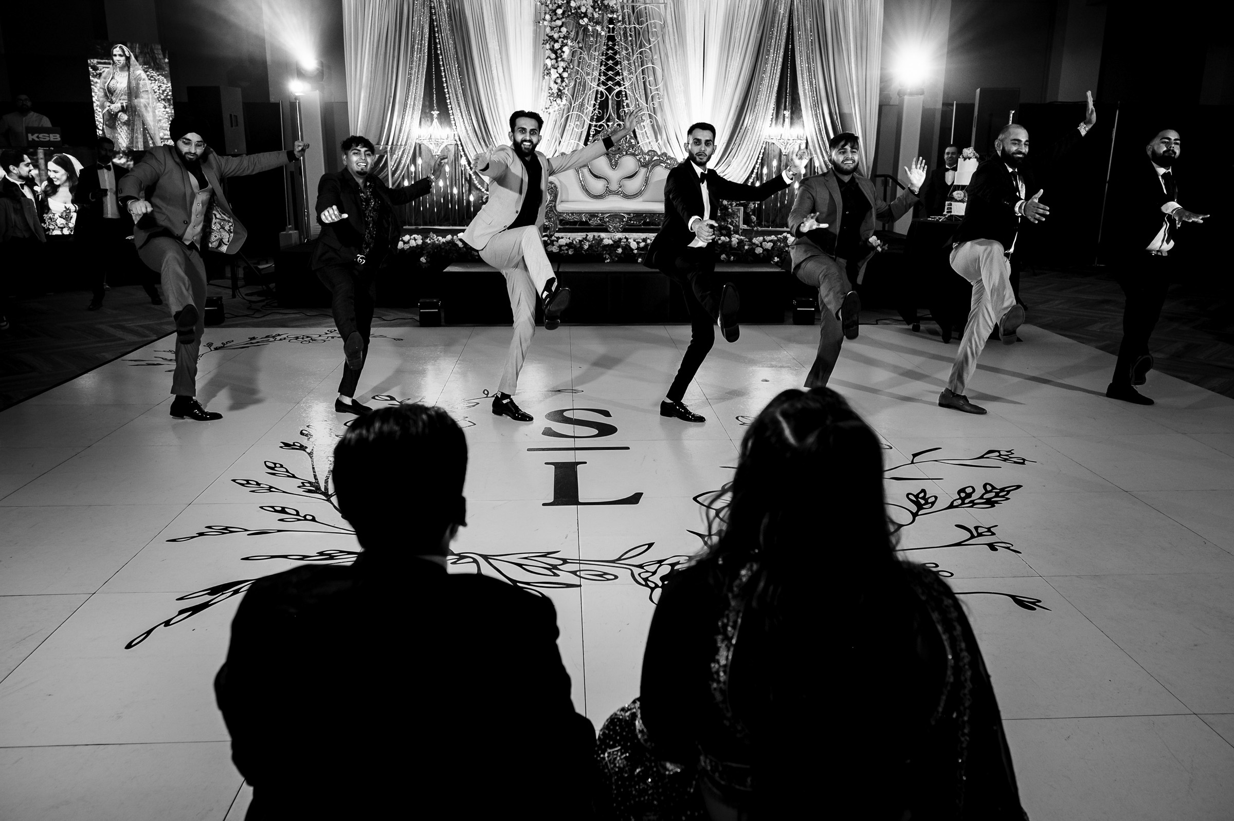 A group of men energetically dancing on a decorated stage, watched by seated guests.