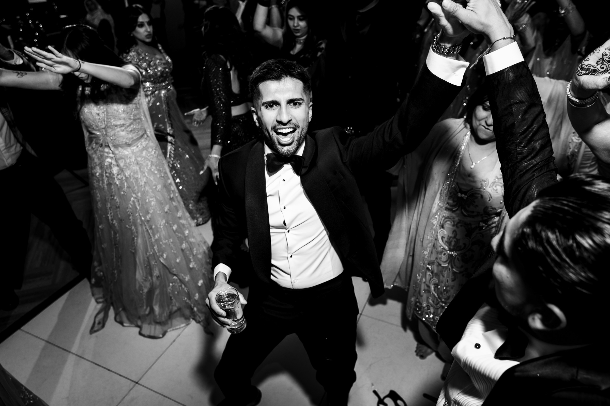 Man in a tuxedo dancing at a lively event, holding a drink as others dance around him.