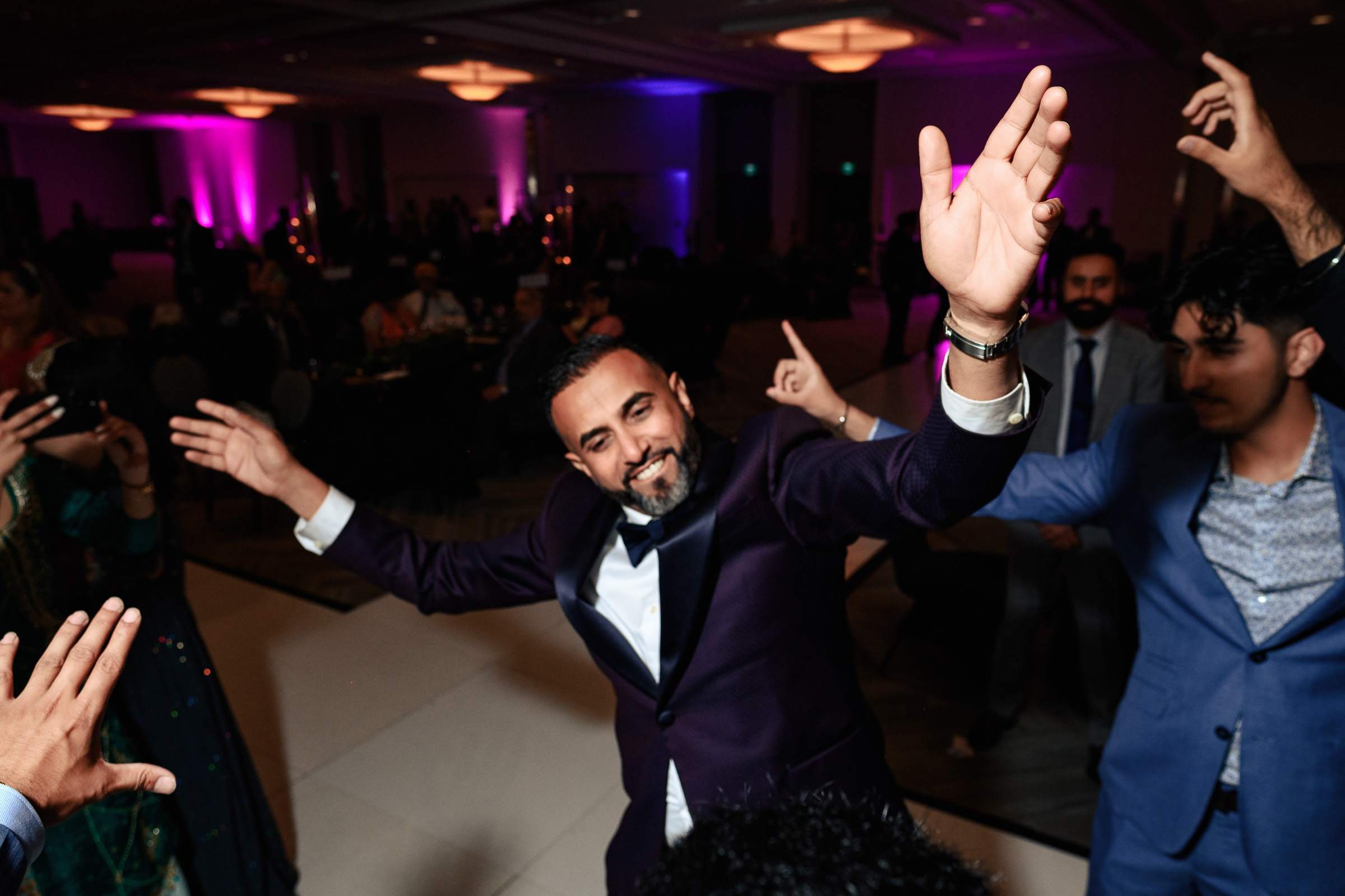 Man in a dark suit dancing energetically at a party with people around.