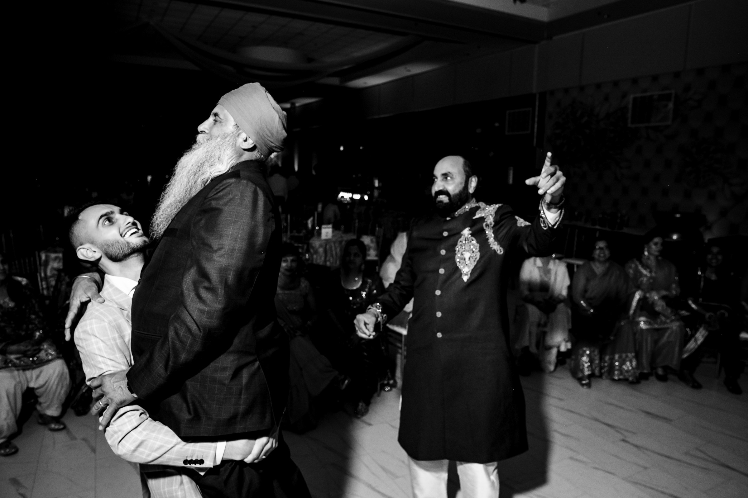 Man lifts another man in celebration while a third man gestures in a lively gathering.