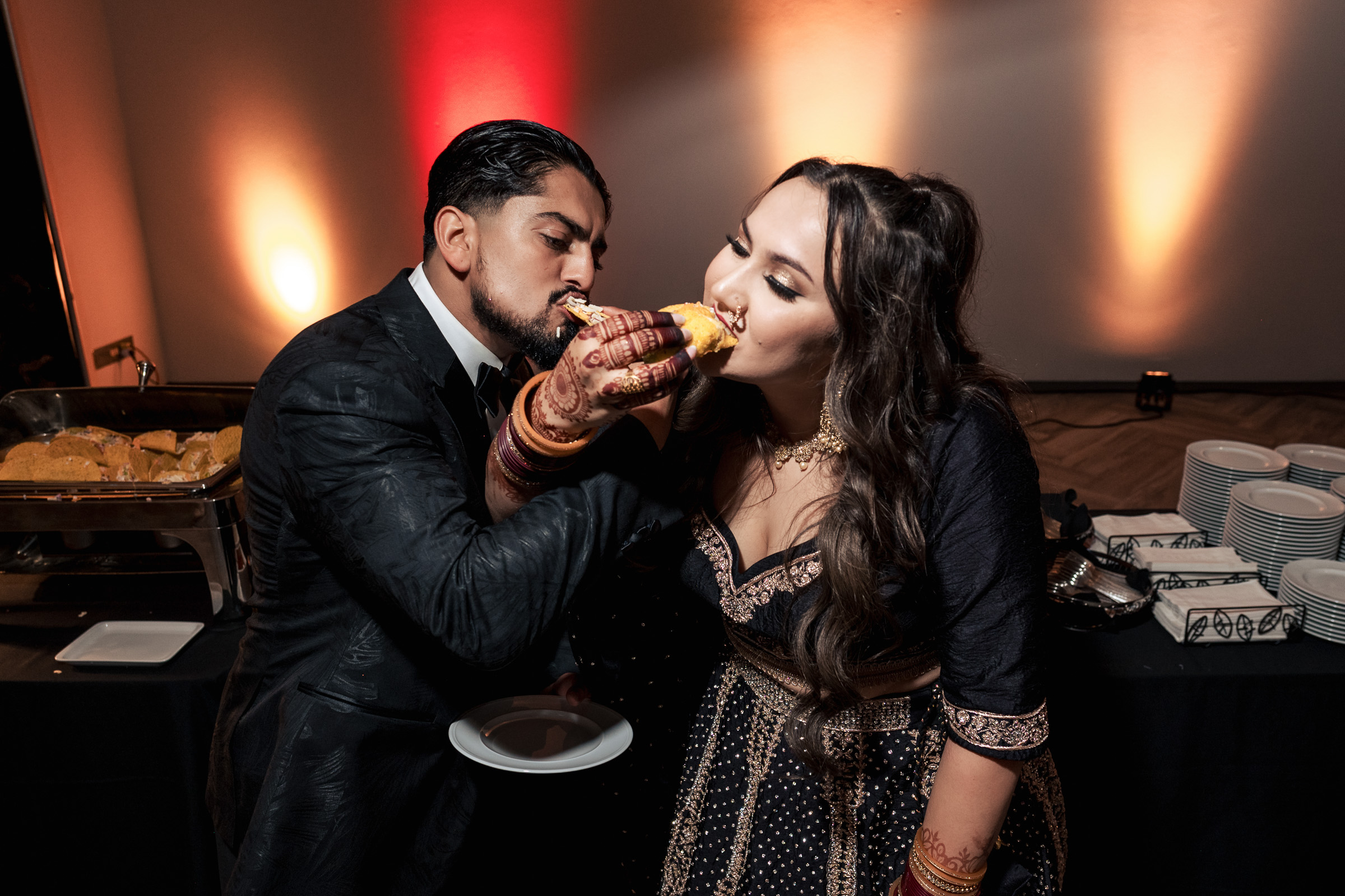 A couple feeds each other a pastry at an indoor event with buffet trays in the background.