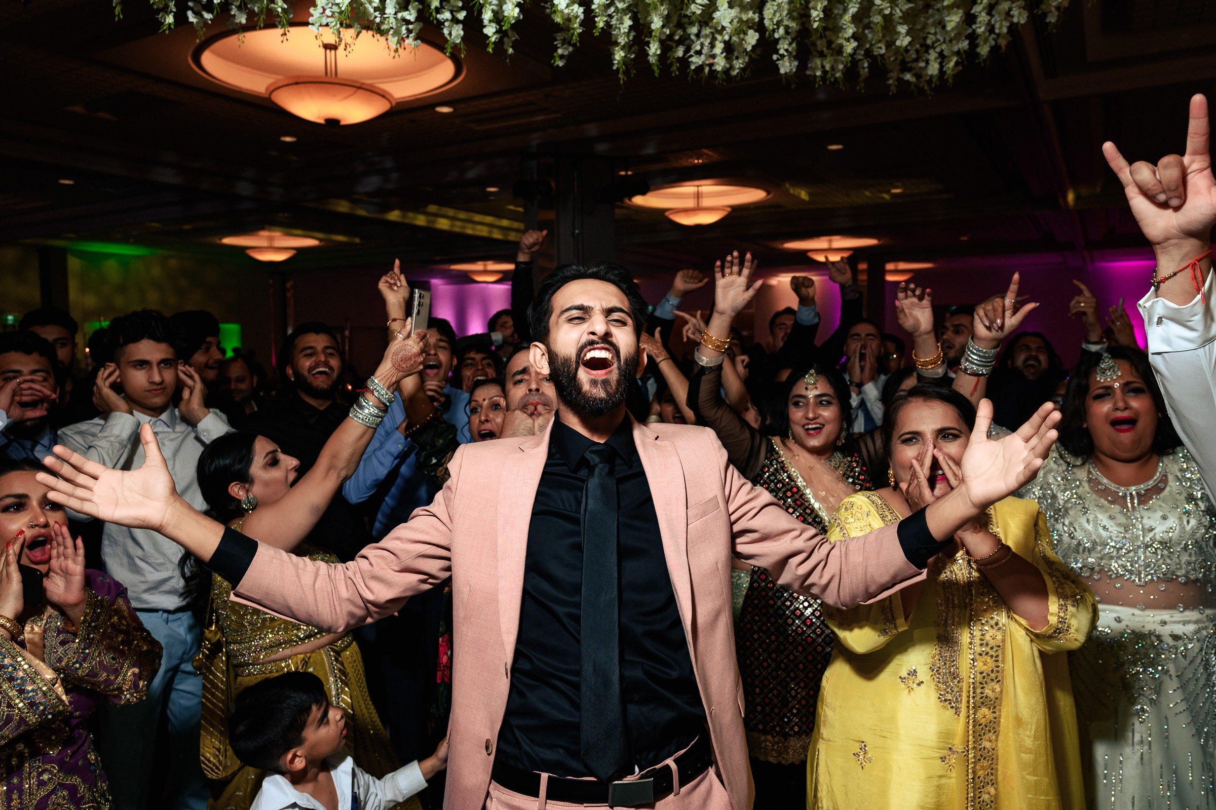 A person in a suit joyfully leads a cheering crowd at a lively indoor event.