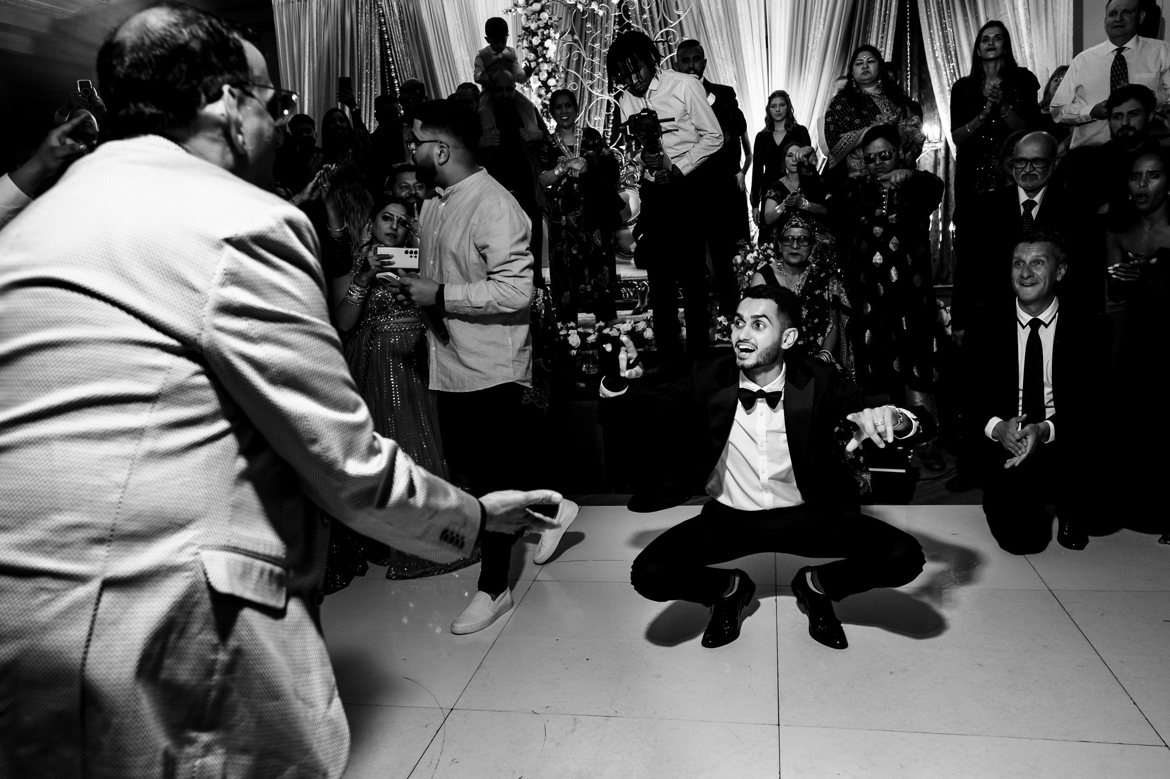 Man crouching energetically on dance floor at lively event with onlookers in formal attire.