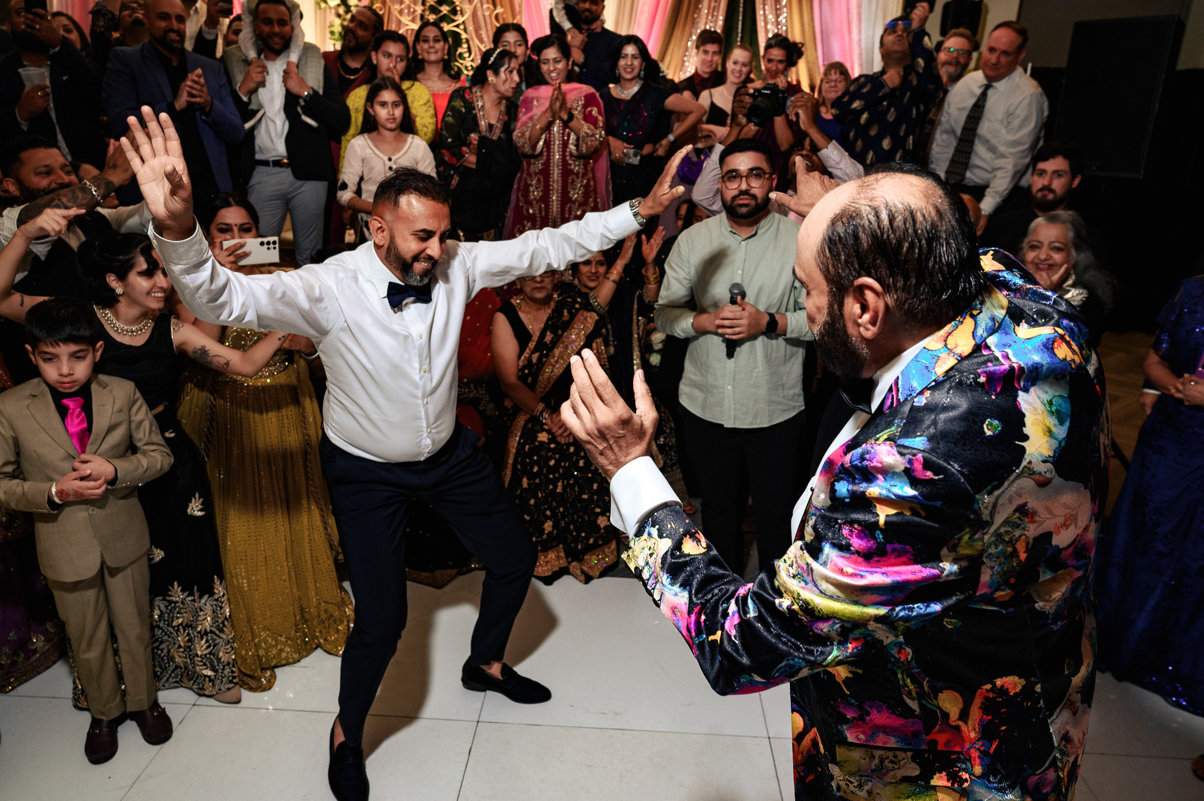 Two men dance energetically at a lively, colorful celebration surrounded by people.