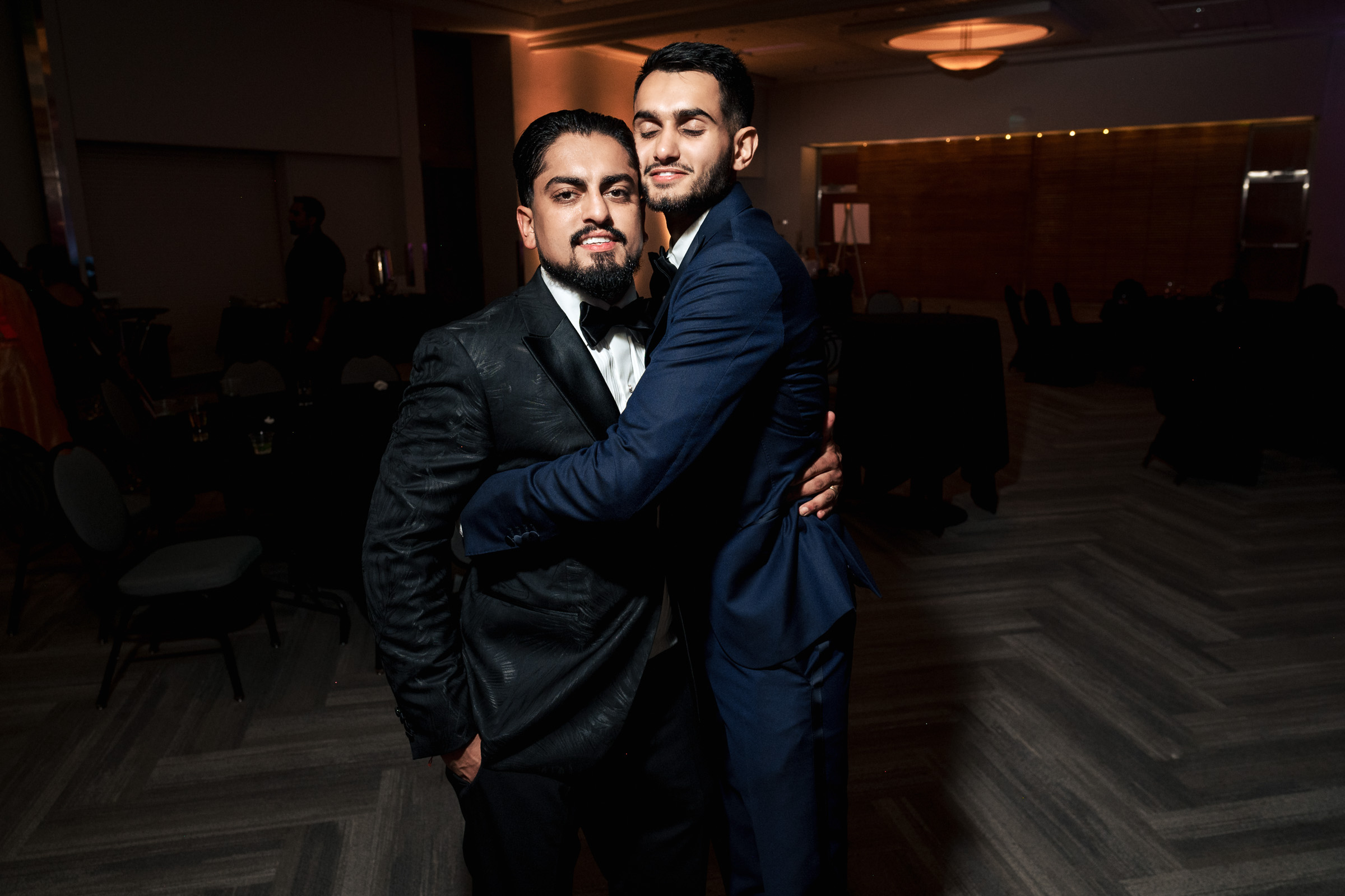 Two men in suits embrace at an indoor event with dim lighting.