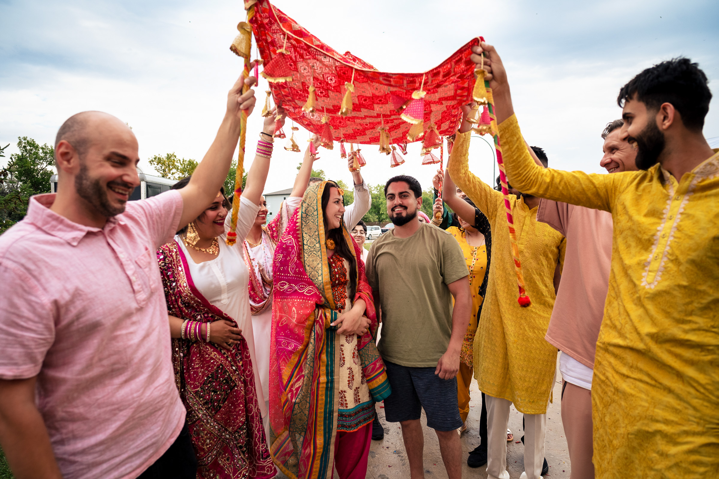 Group of people holding a red decorative cloth over a smiling couple outdoors.