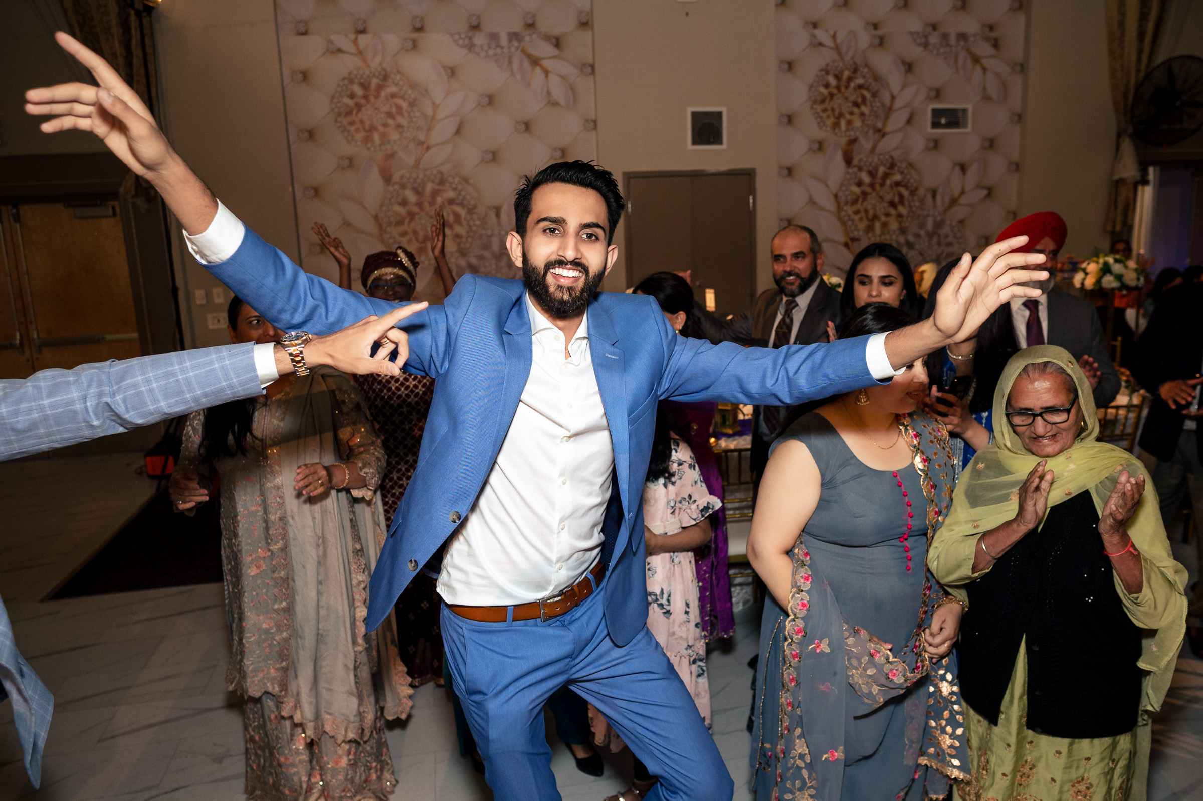 Man in blue suit joyfully dancing at a lively indoor gathering.