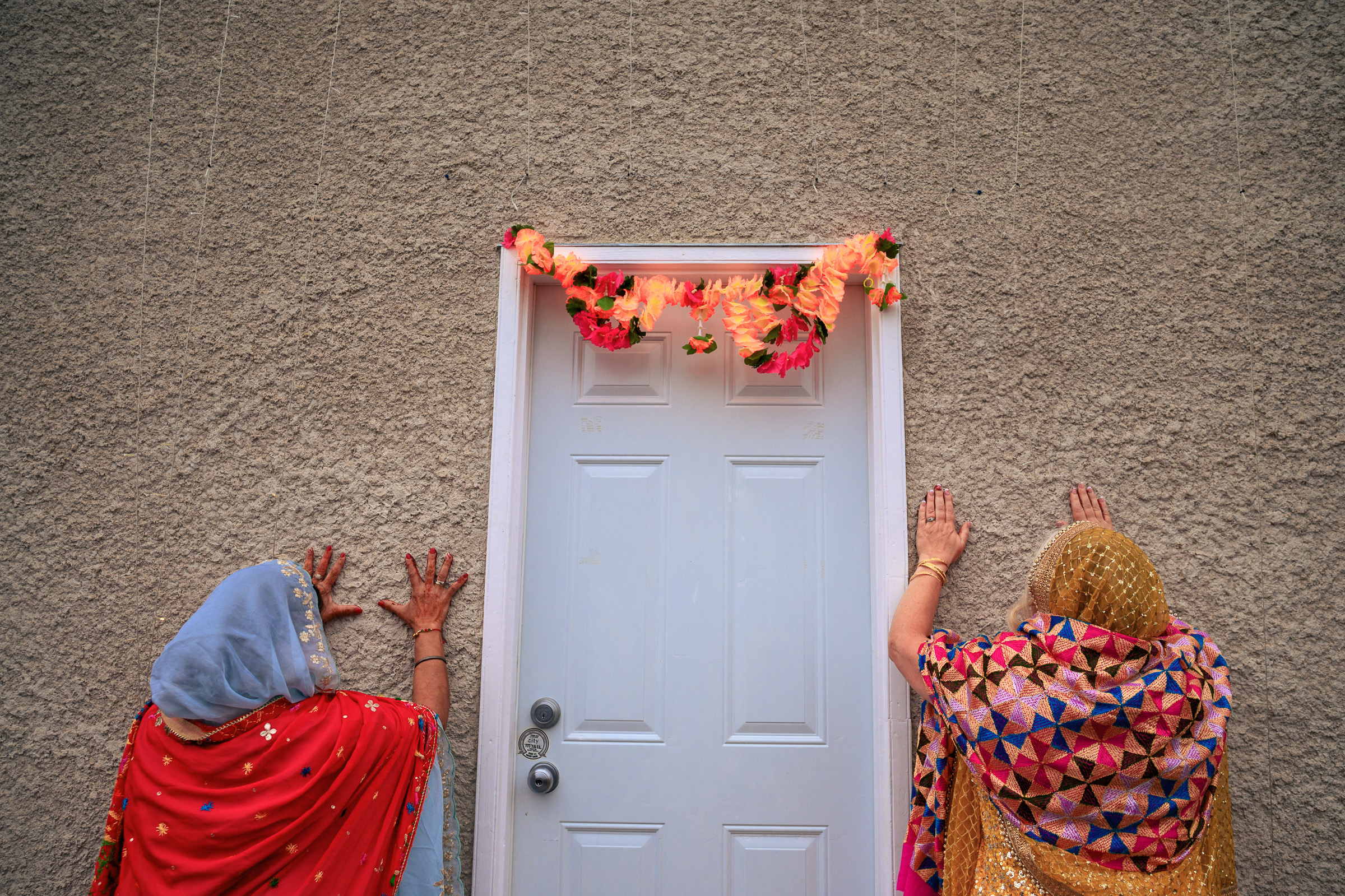 Two women in colorful attire stand facing a decorated white door with their hands on the wall.
