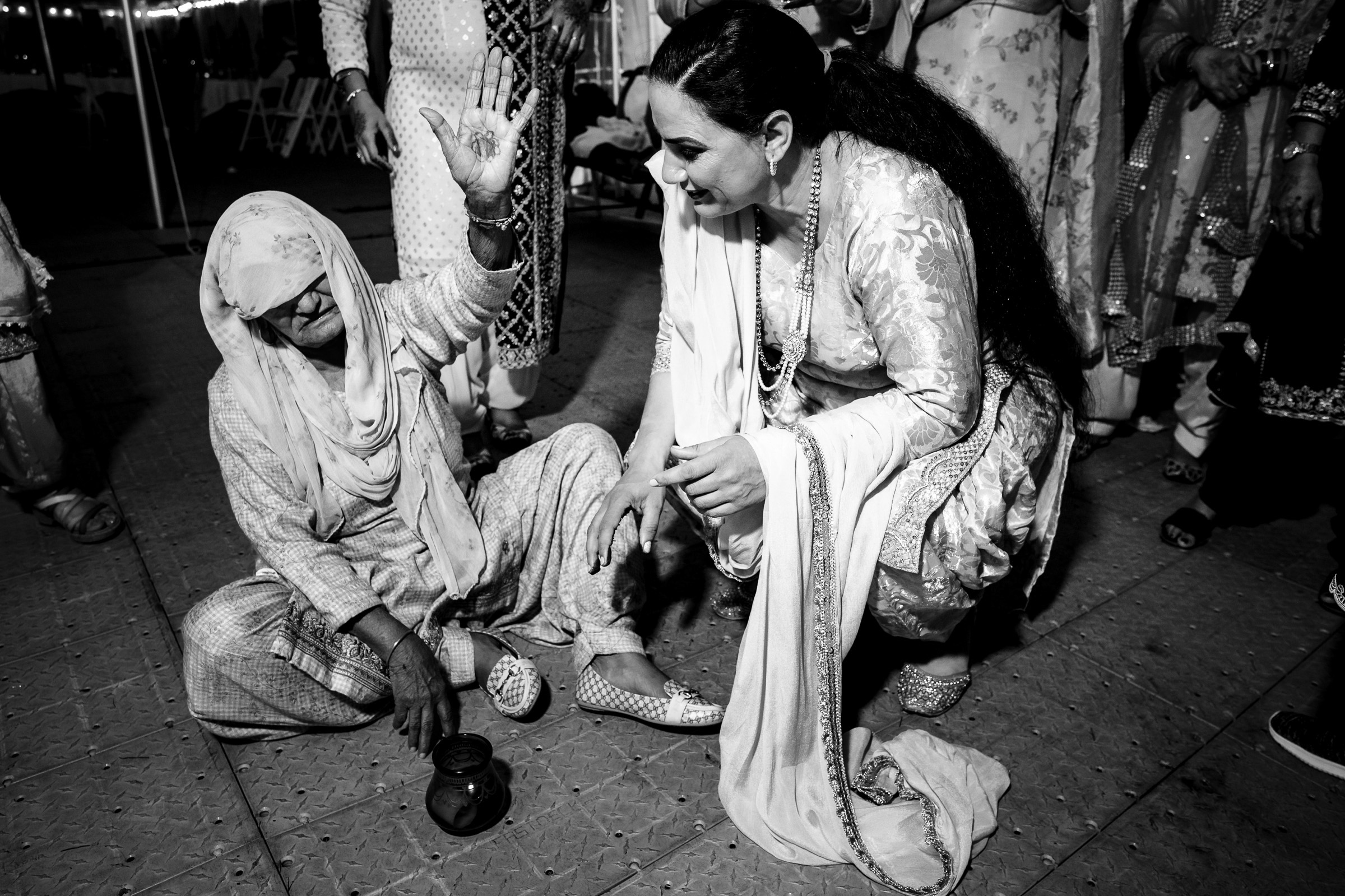 Two women in traditional attire engage in conversation; one is seated, holding a small object.