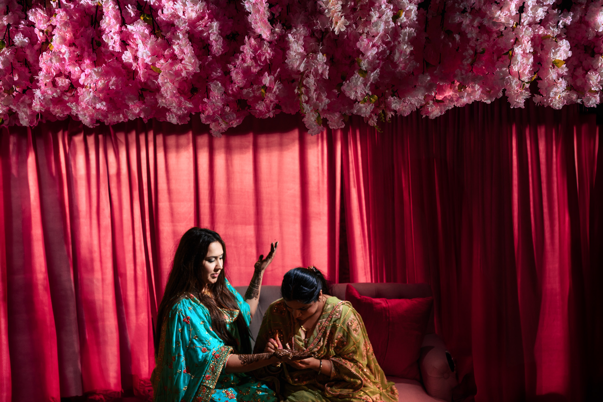 Two women in colorful attire under pink flower decorations with red curtains in the background.