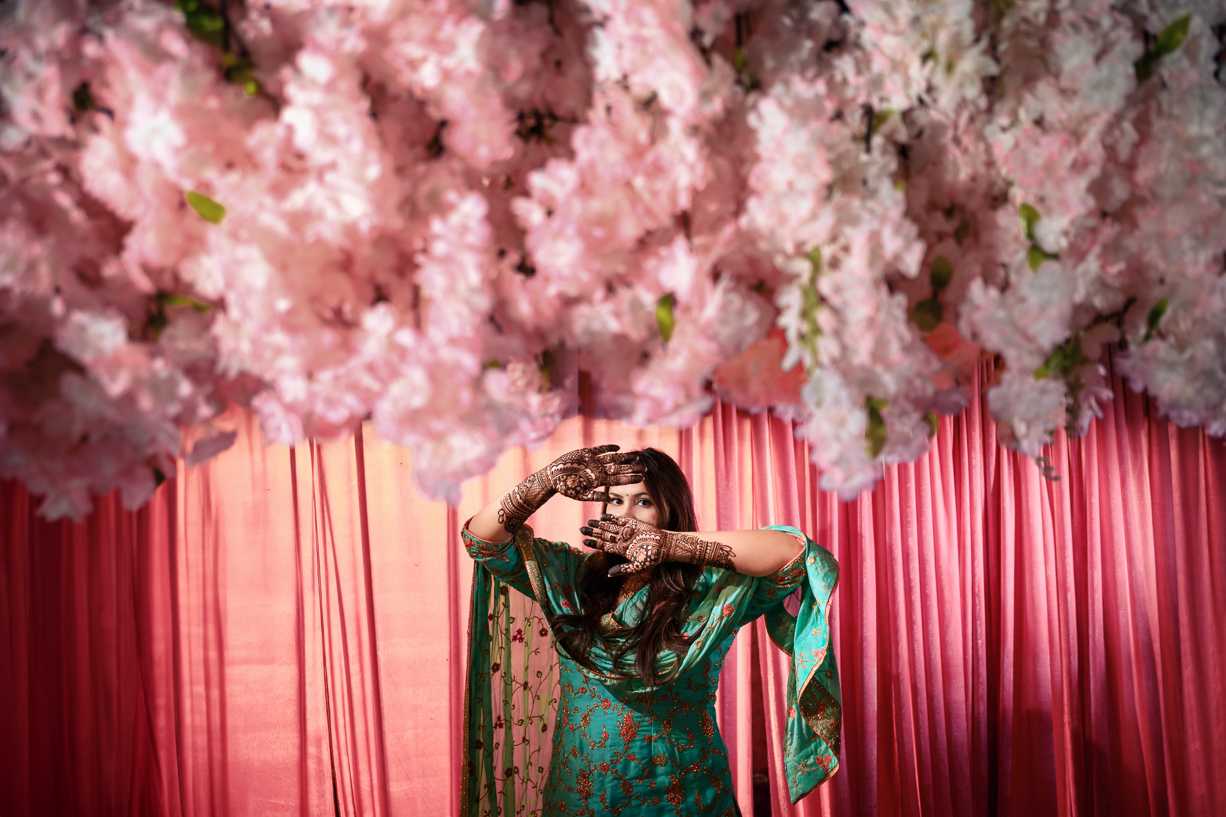 Person in green attire poses under pink floral decoration with hennaed hands raised.