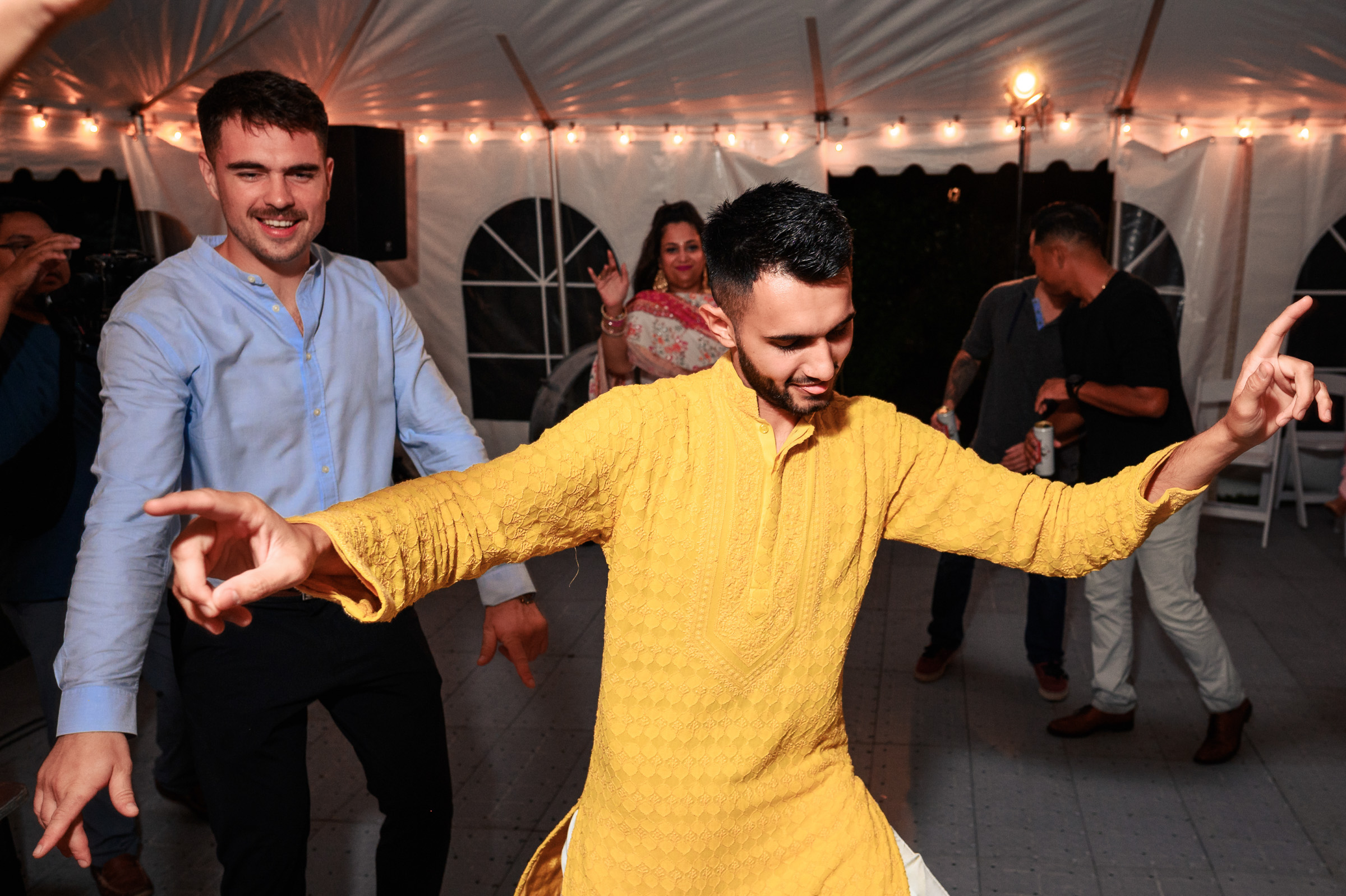 People dancing energetically under a tent with string lights.