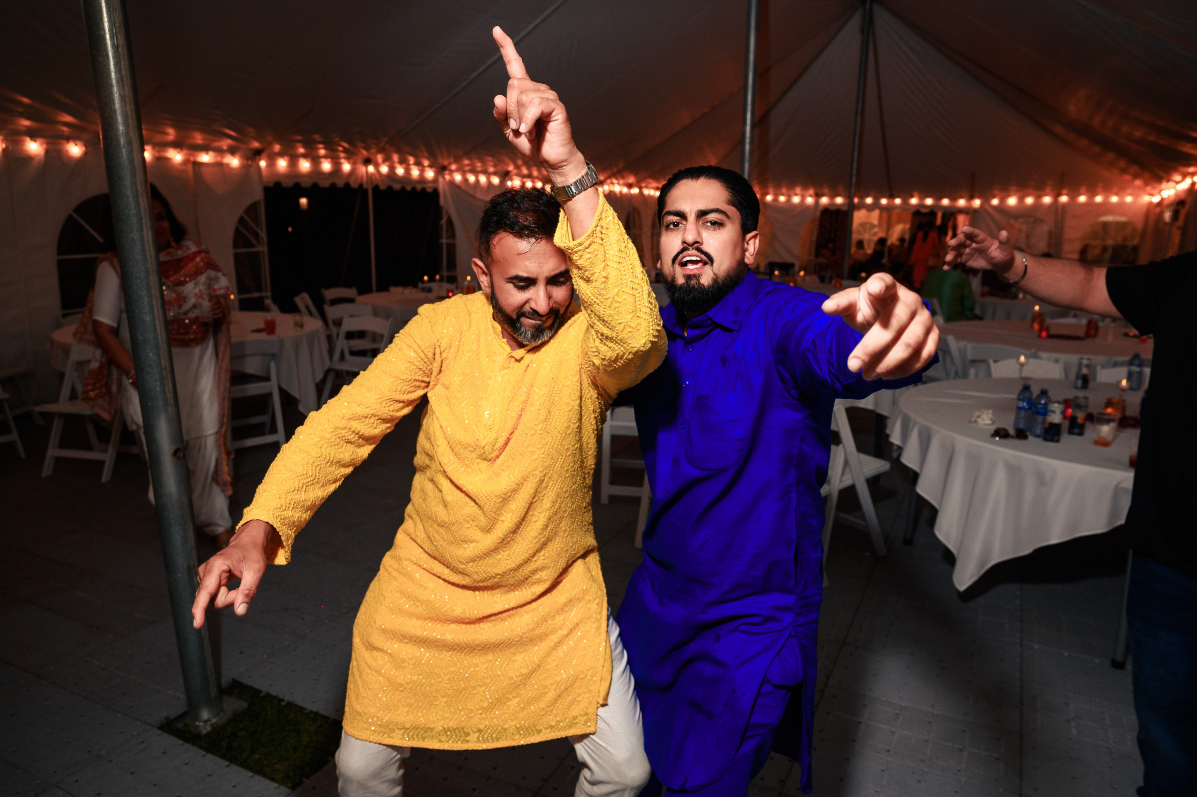 Two men in colorful traditional attire dancing enthusiastically at an event under a tent.