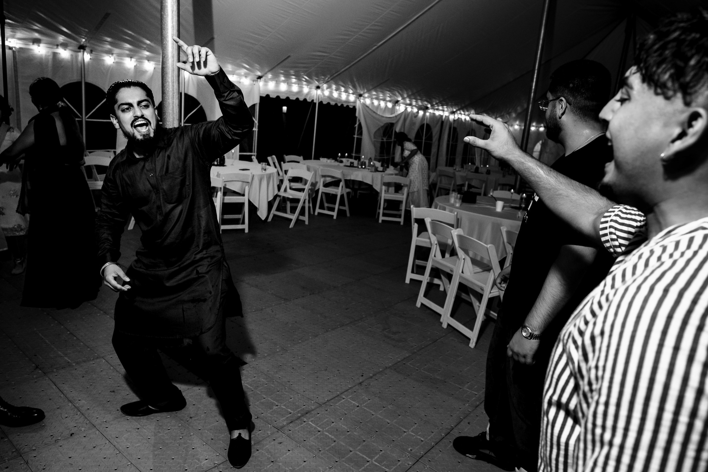People dancing energetically under a lit tent with empty tables in the background.