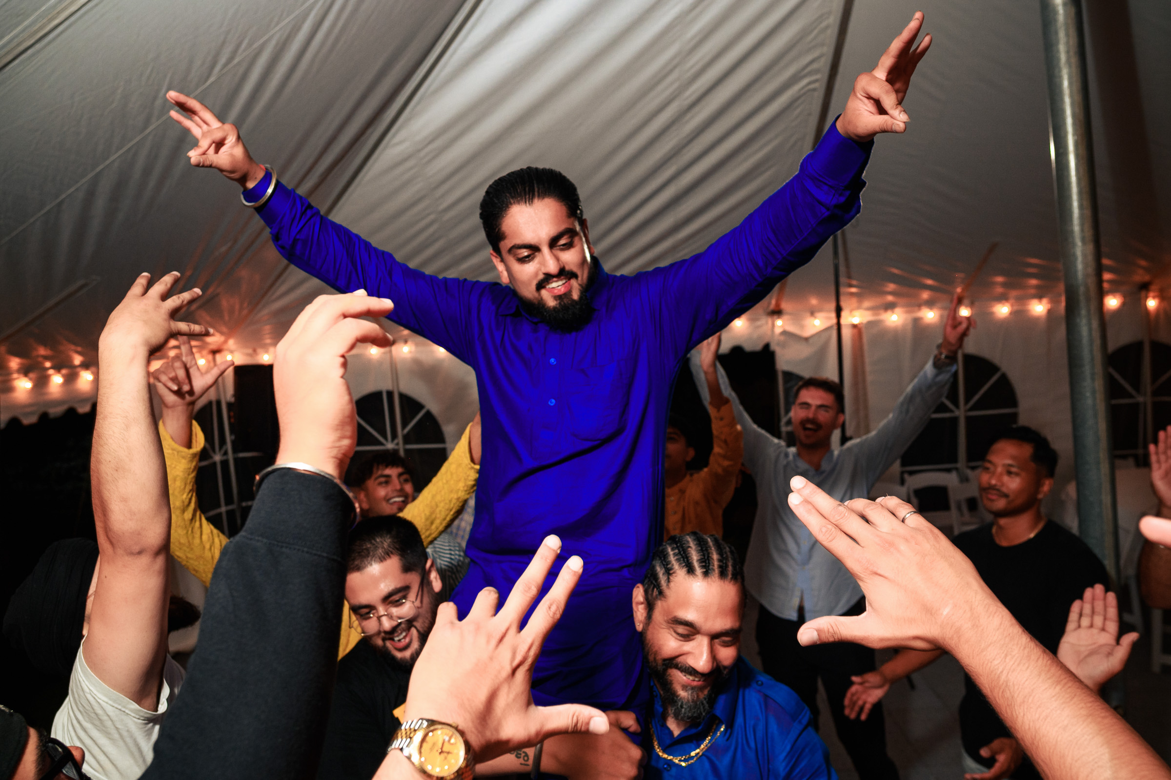 A man in a blue outfit is lifted up by people at a lively celebration under a tent.