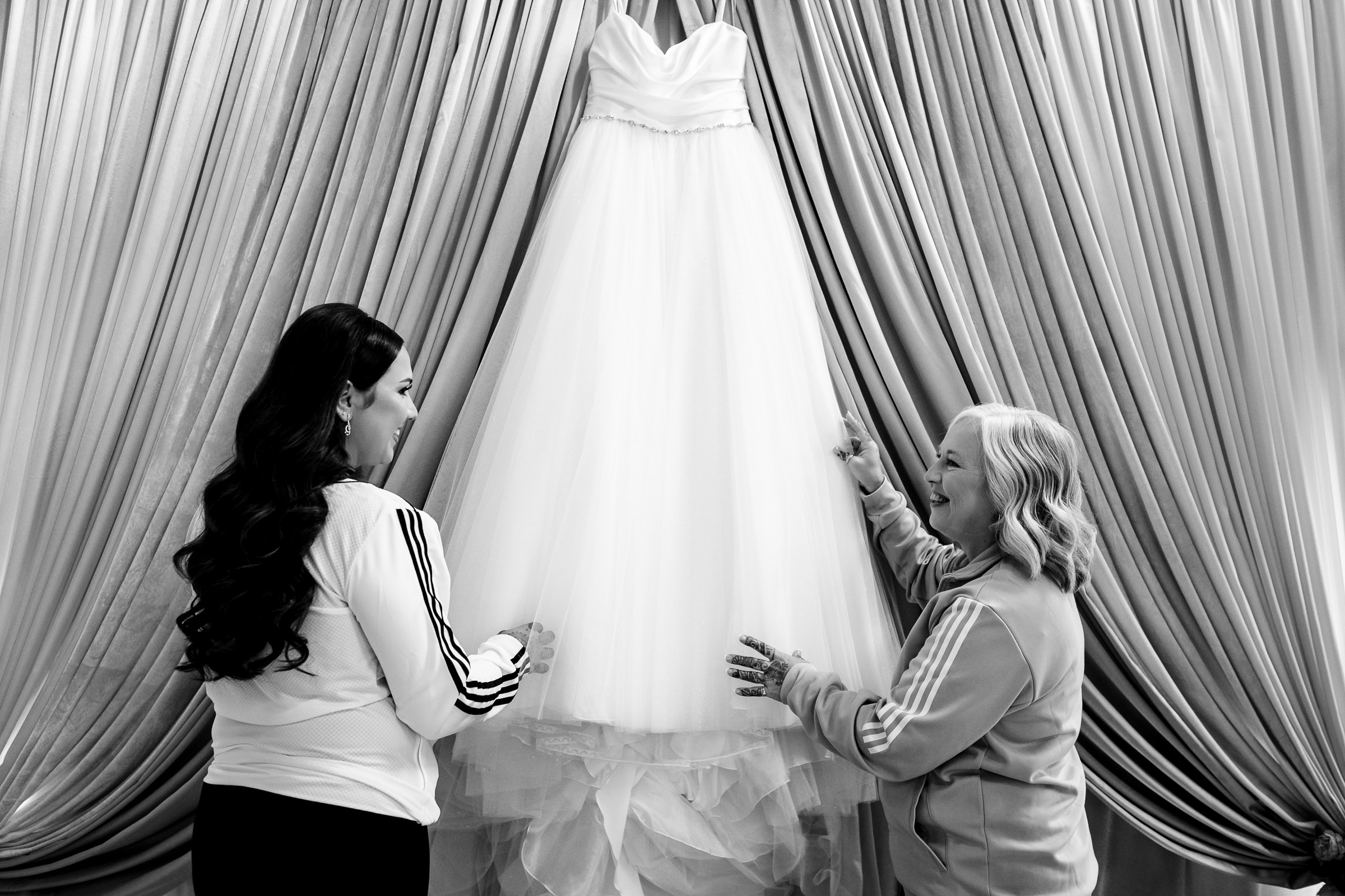 Two women smile and gesture toward a hanging wedding dress against draped curtains.