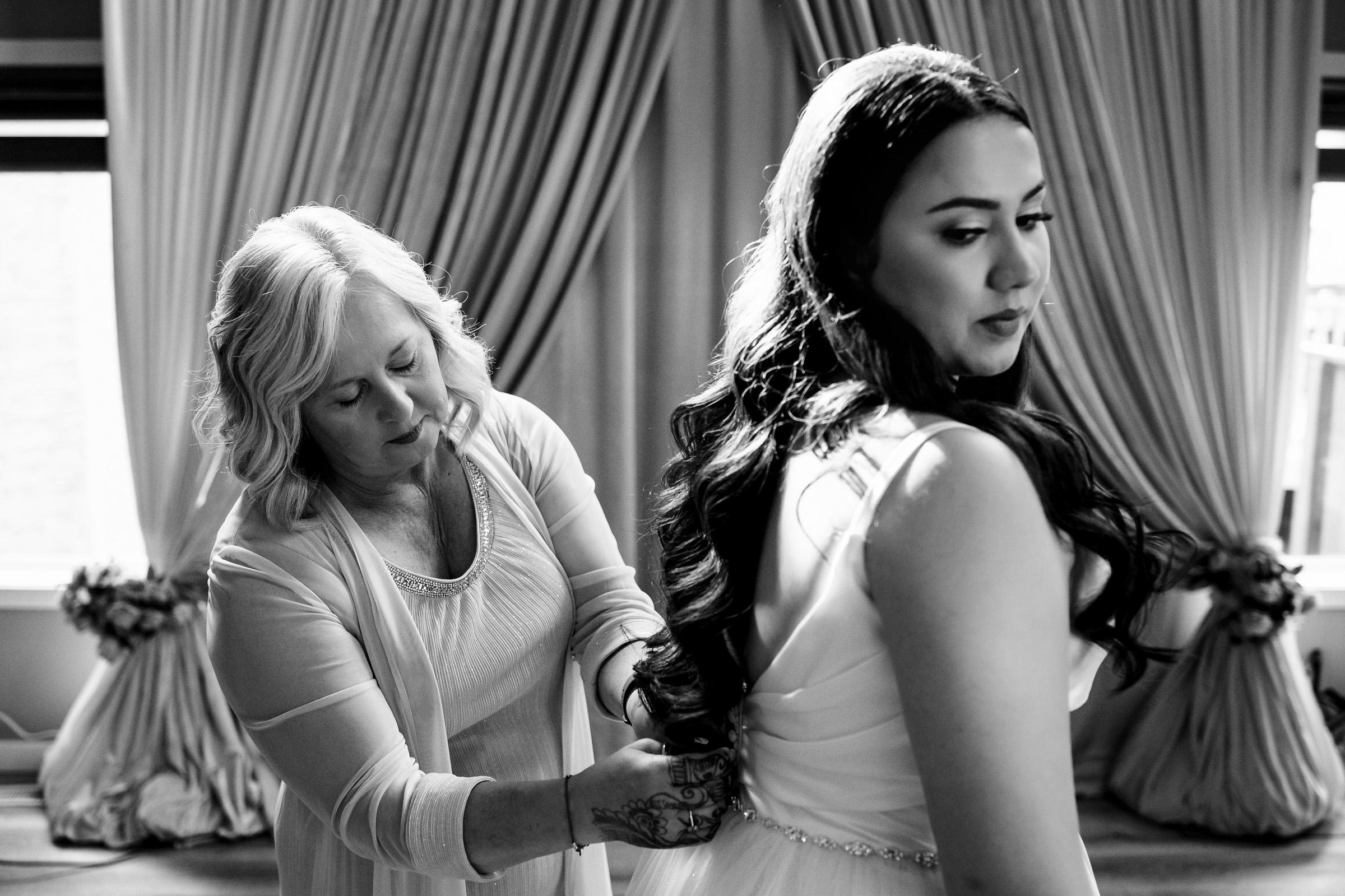 Woman helps another adjust her dress in a room with draped curtains.
