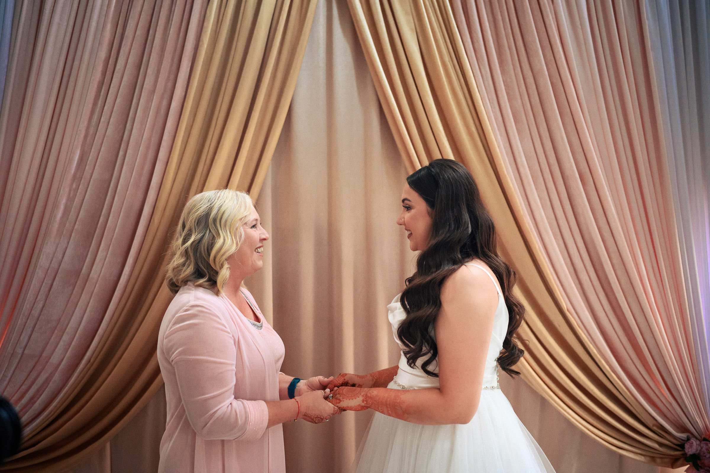 Two women smiling at each other, holding hands in front of draped curtains.