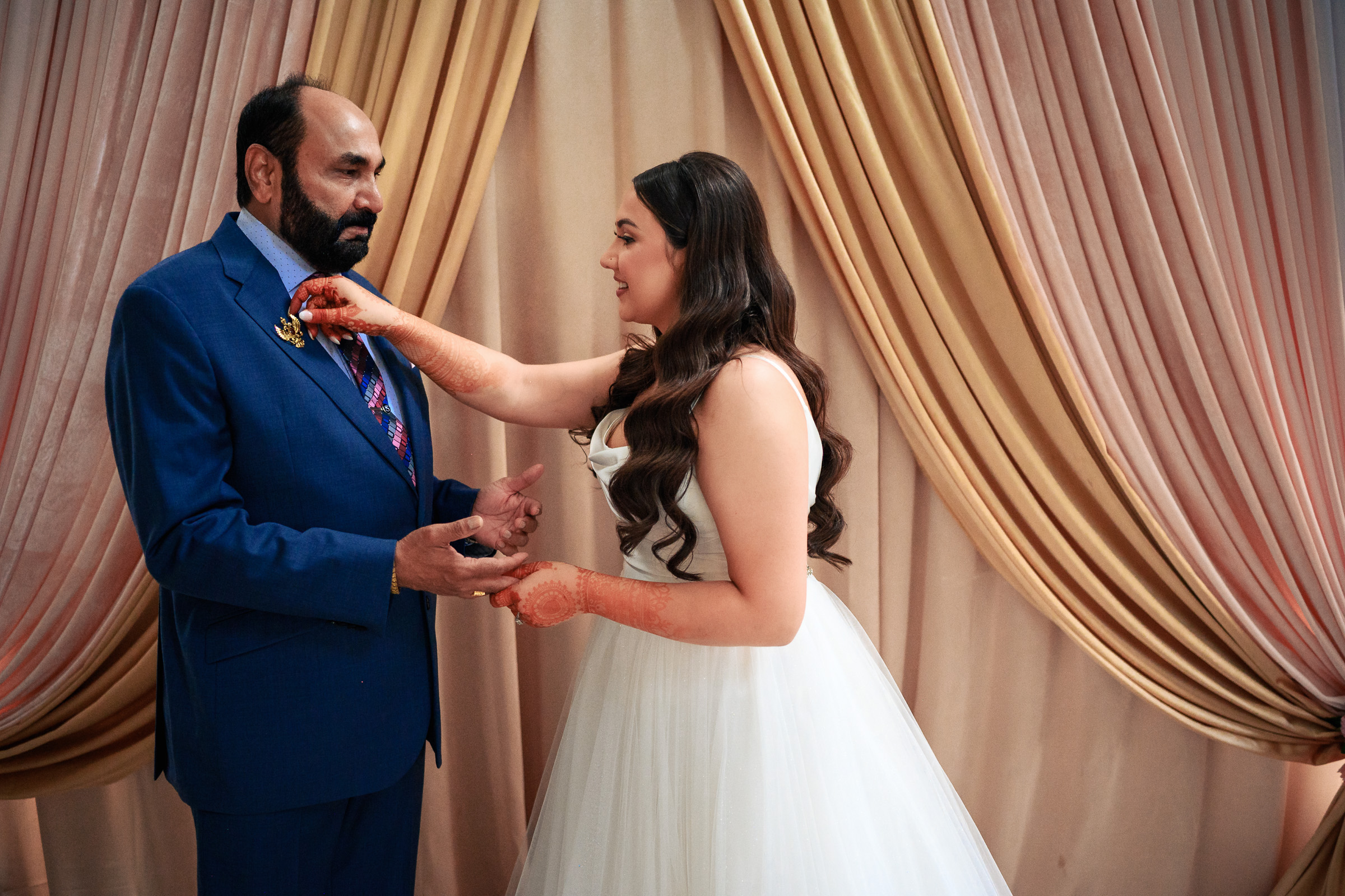 Bride adjusts man's suit in front of gold and pink drapes.
