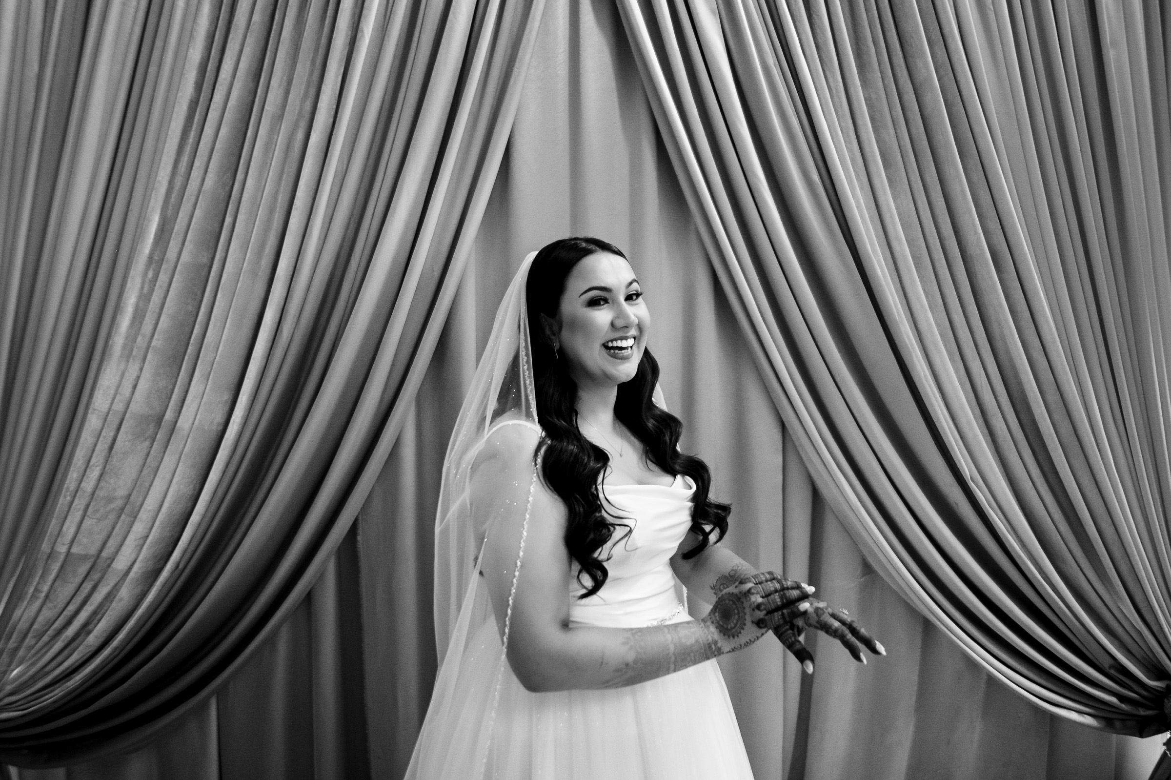 Bride smiling with henna on hands, standing in front of draped curtains.