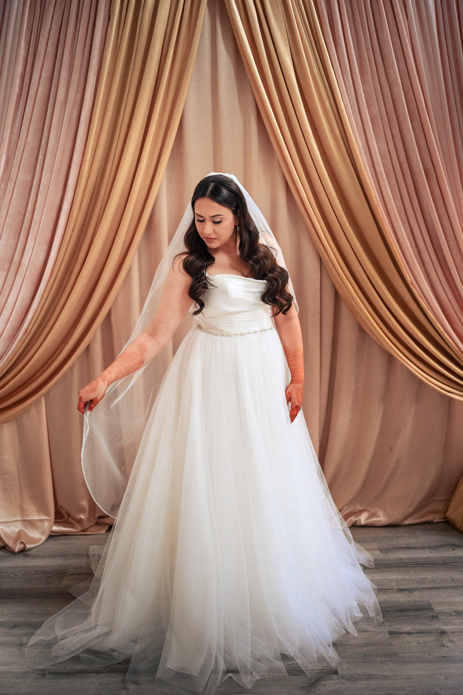 Bride in white gown with henna on hands, standing before gold draped curtains.