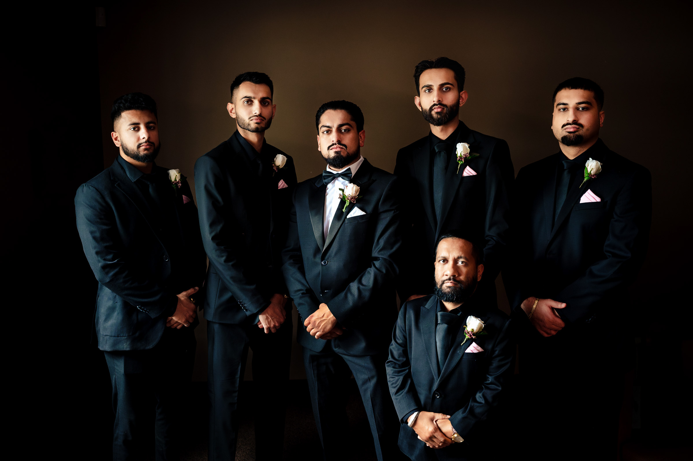 Six men in black suits with boutonnieres stand against a dark background.