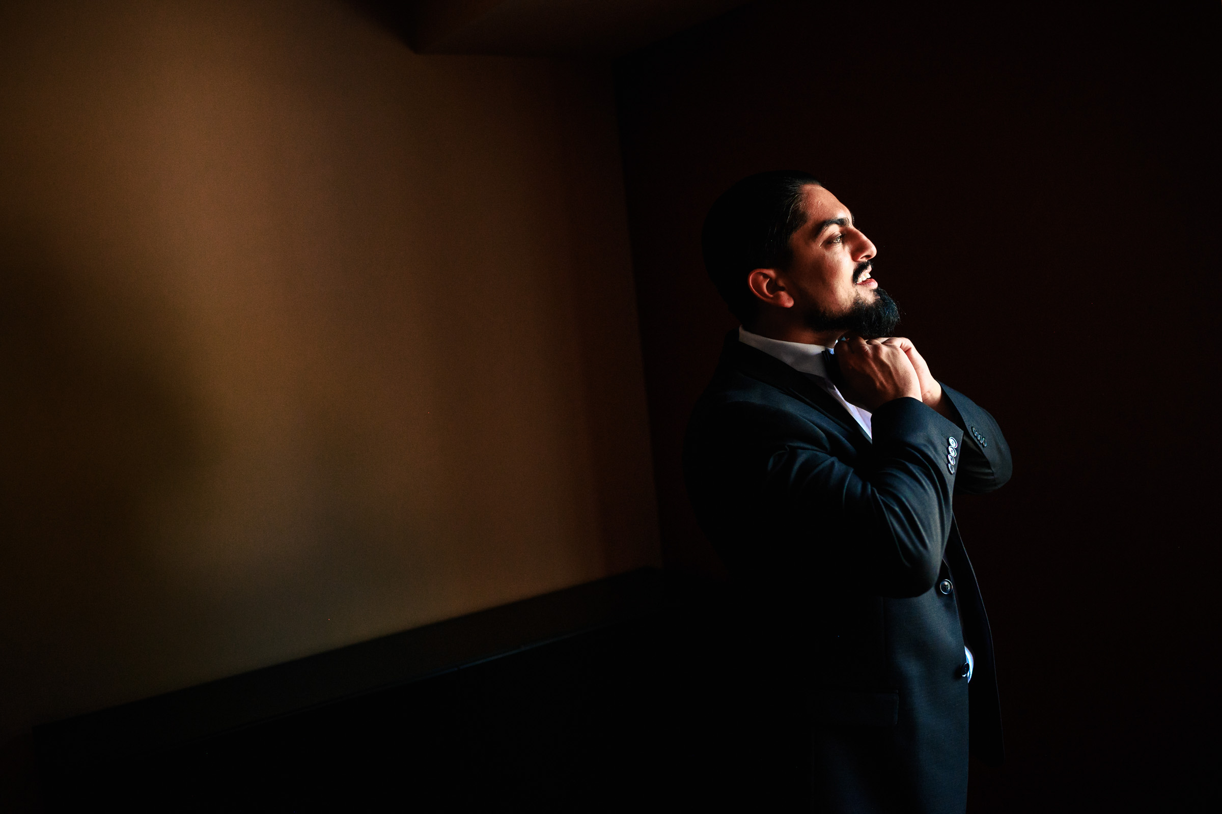 Man adjusting his bow tie, standing in a dimly lit room with a dark background.