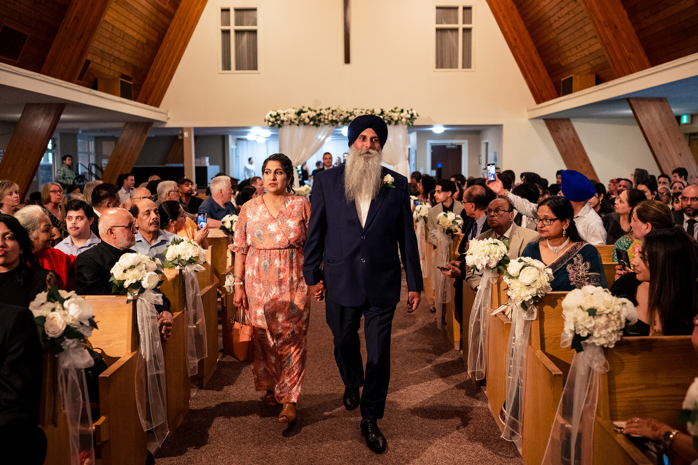 Two people walking down the aisle in a church with seated guests on either side.