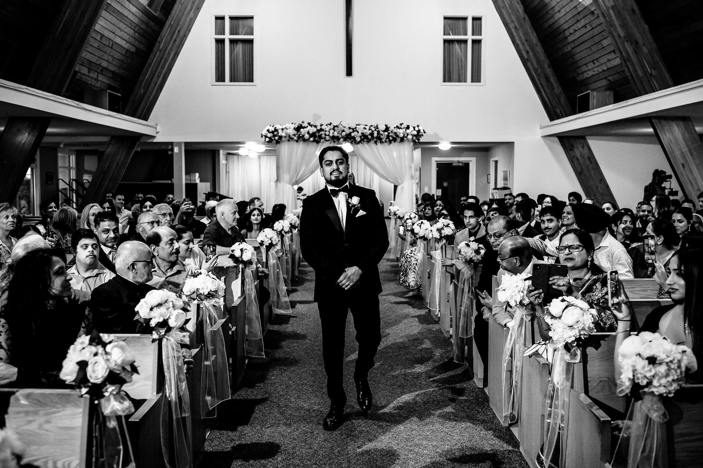 Groom walks down the aisle in a church, surrounded by seated guests.