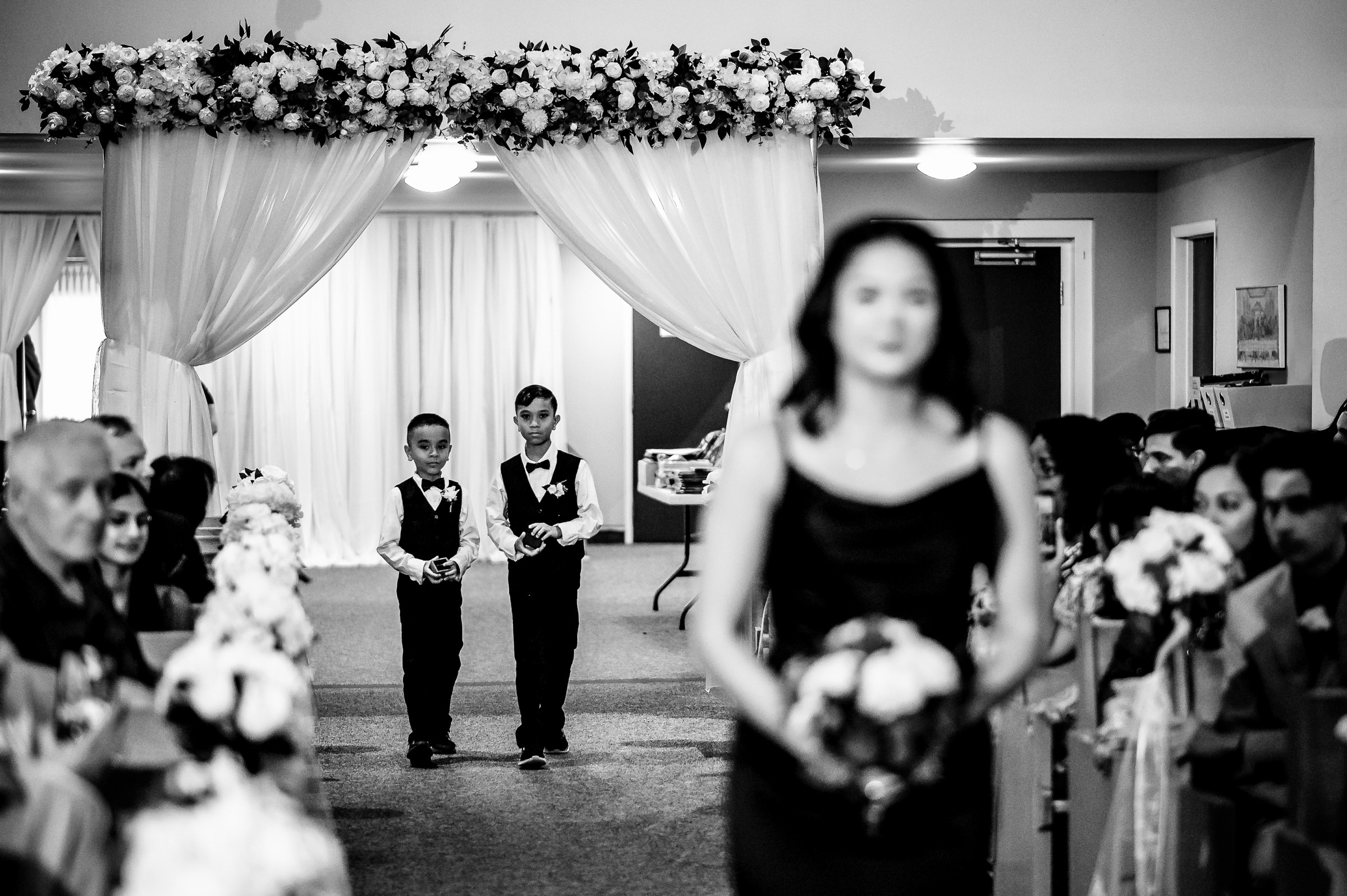 Two boys walk down an aisle at a wedding ceremony, carrying small items.