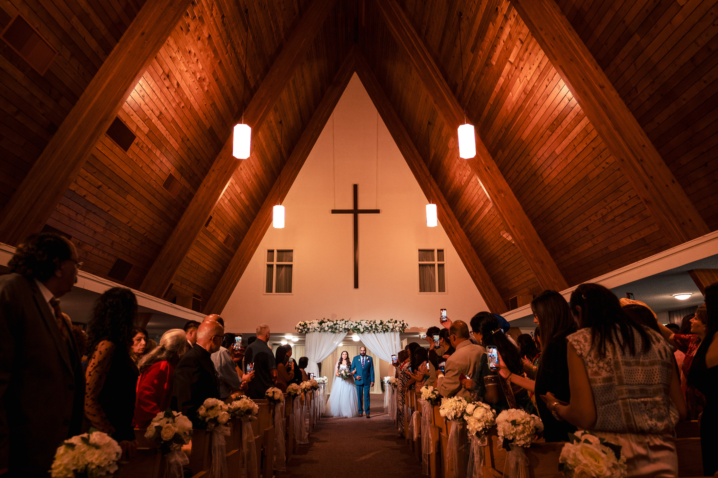 Bride and groom walk down the aisle in a wooden church, guests stand and take photos.